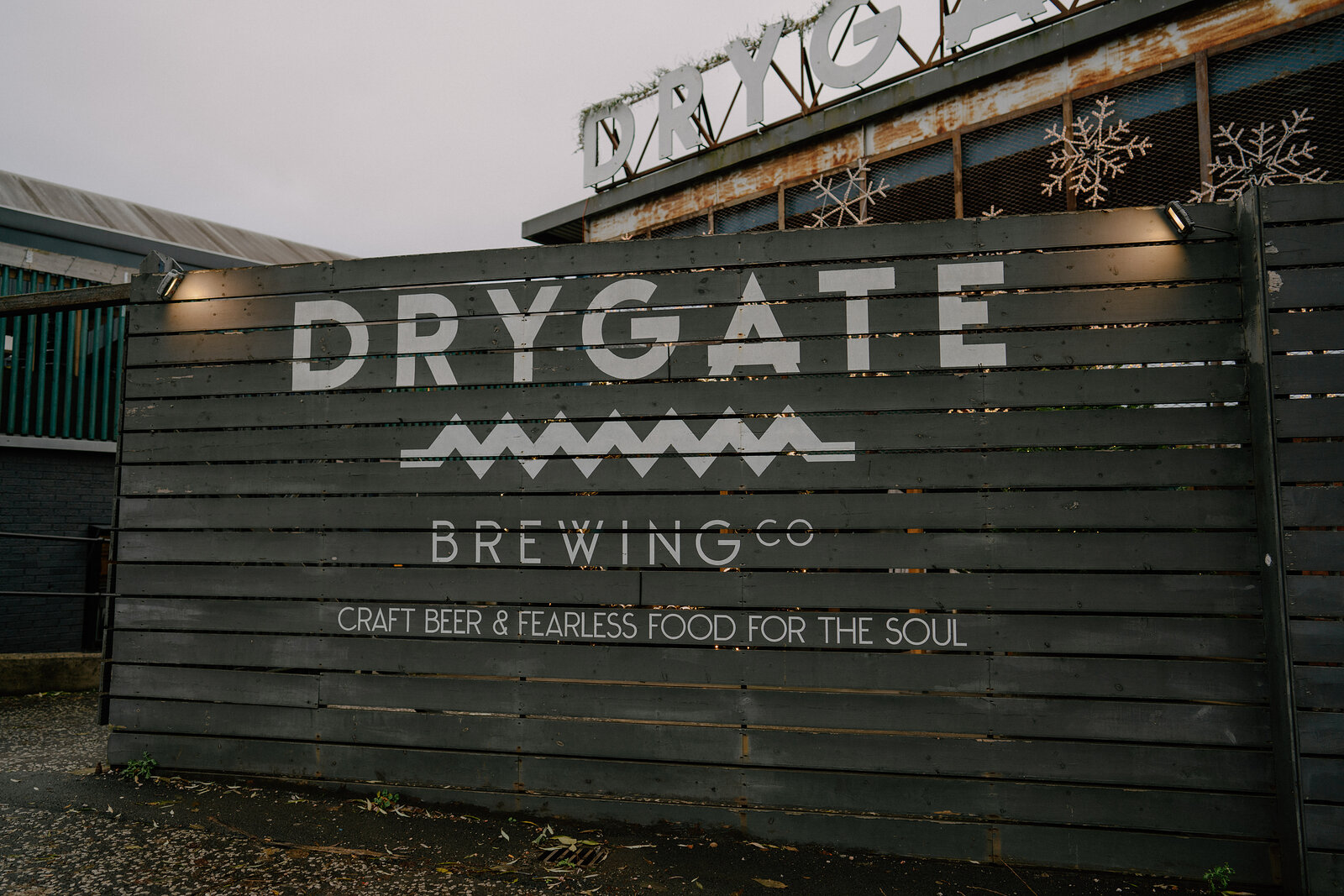 A mural on dark timber cladding reads "DRYGATE BREWING Co — CRAFT BEER & FEARLESS FOOD FOR THE SOUL", snowflake lights strung along the roofline above.