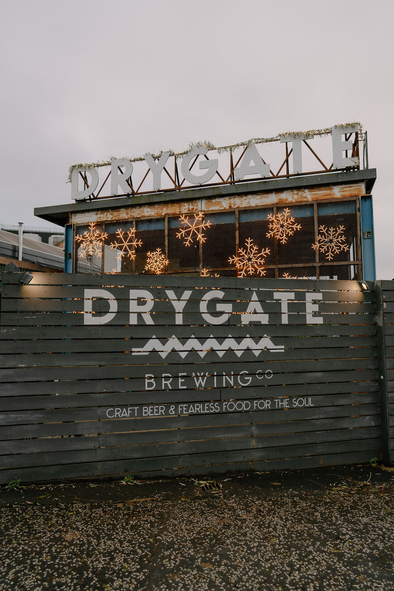 A mural reading "DRYGATE BREWING Co — CRAFT BEER & FEARLESS FOOD FOR THE SOUL" is trimmed with a tinsel garland along its rooftop lettering, snowflake lights glowing in the windows below.