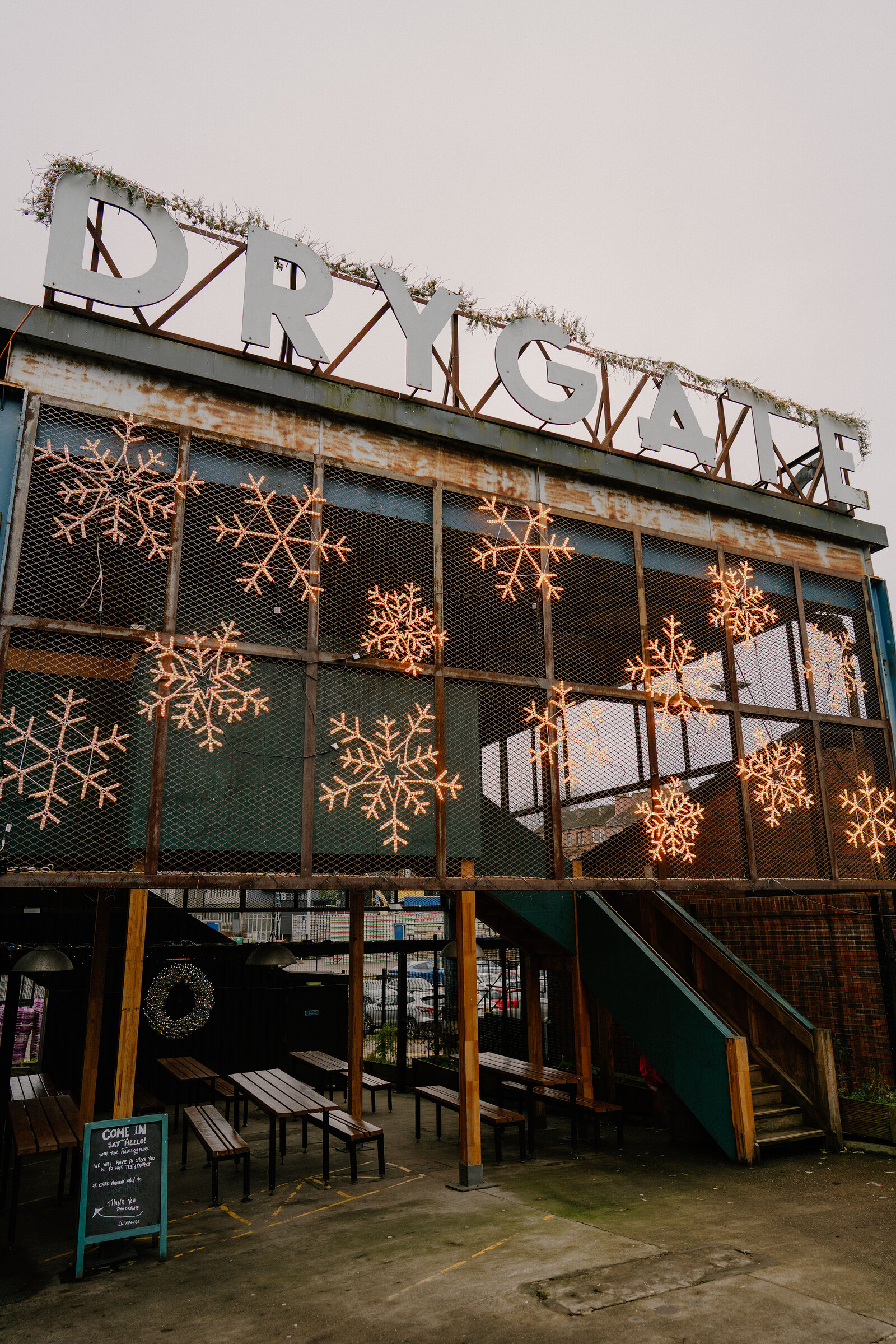 Illuminated snowflakes cover a mesh screen beneath rooftop lettering reading "DRYGATE" trimmed with tinsel, picnic benches and a chalkboard reading "Come In" set on the terrace below.