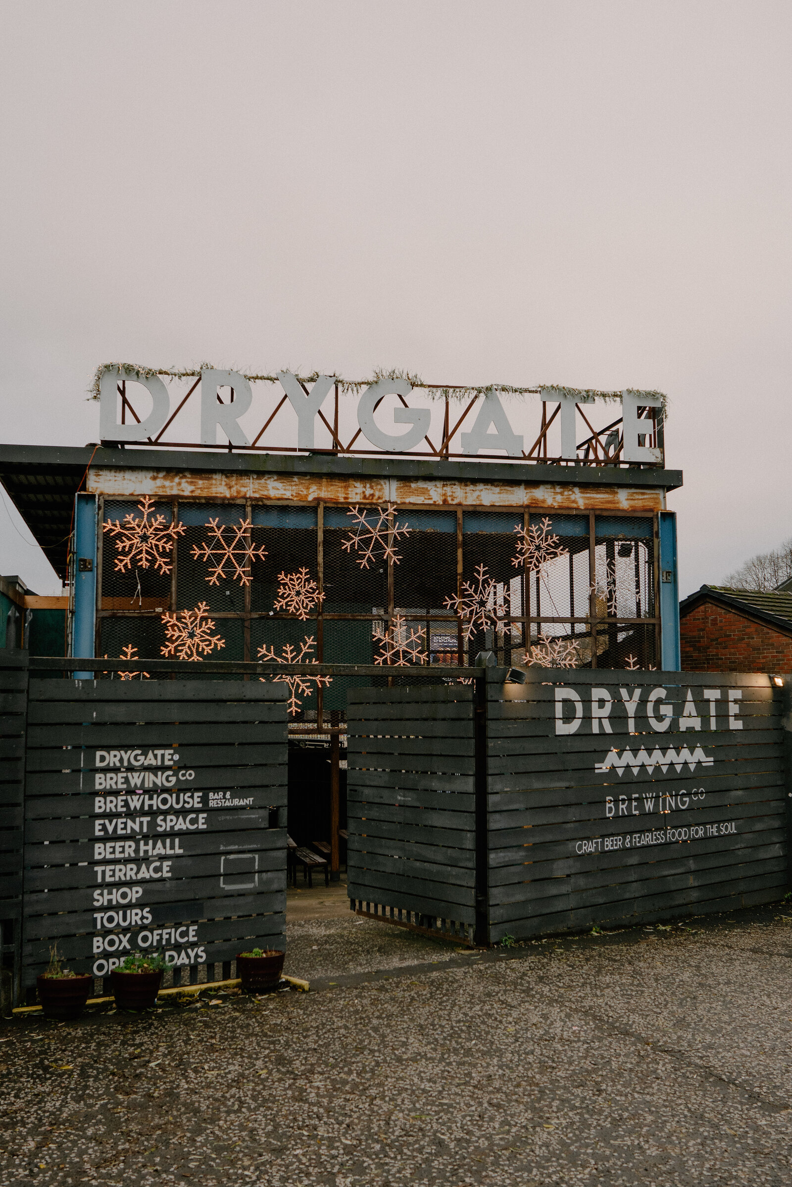 A signboard reading "DRYGATE BREWING Co — BREWHOUSE, EVENT SPACE, BEER HALL, TERRACE, SHOP, TOURS, BOX OFFICE, OPEN 7 DAYS" stands beside a gate hung with illuminated snowflakes.