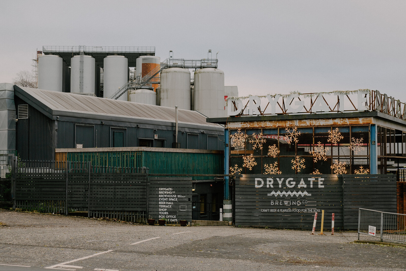 Steel fermentation tanks rise above a corrugated roof beside a mural reading "DRYGATE BREWING" lit with snowflake lights.