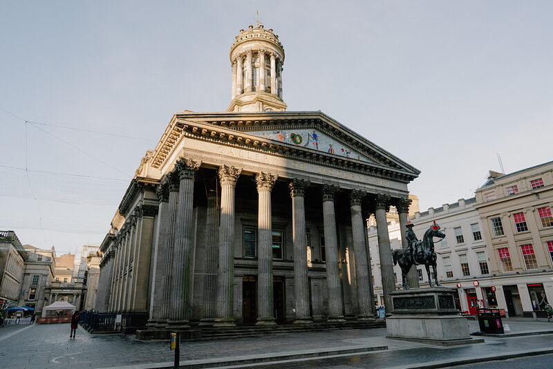 A classical domed building with a colonnade of tall columns rises on a street corner, an equestrian statue standing in front.