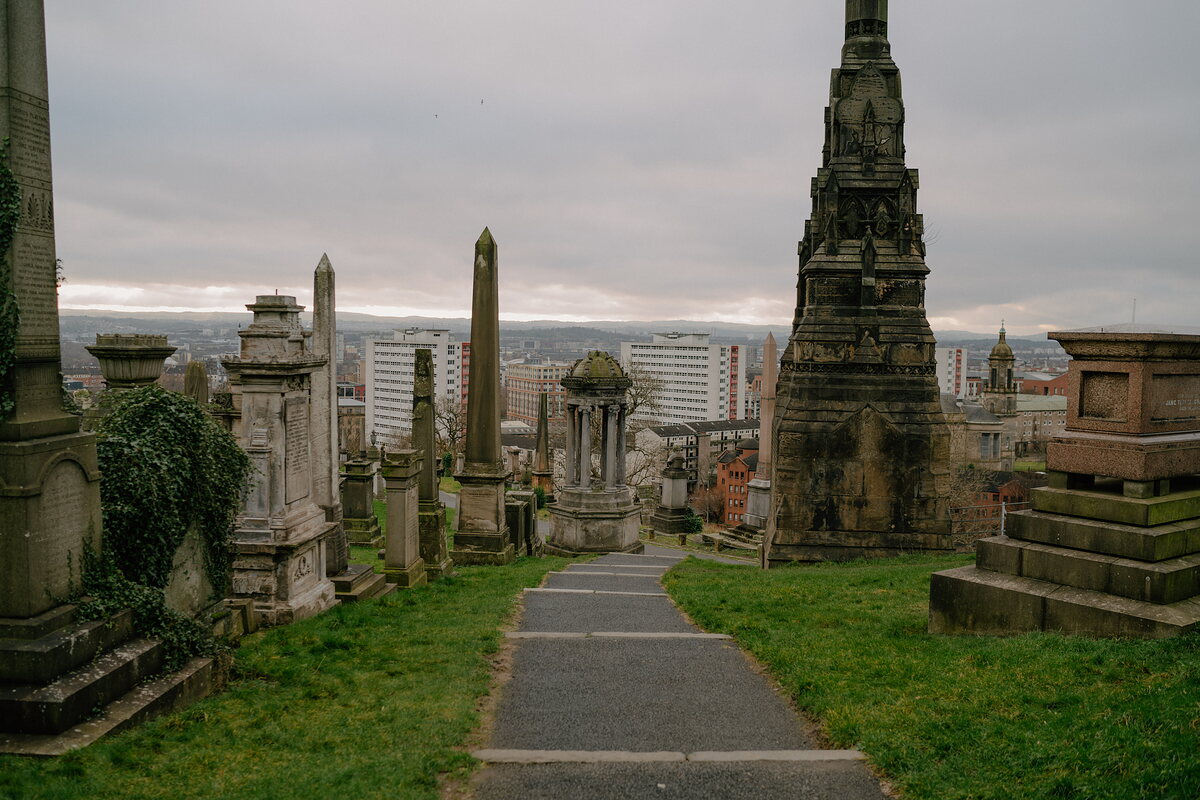 A path winds downhill between tall obelisks and Victorian monuments in a hillside cemetery, city tower blocks visible in the distance.