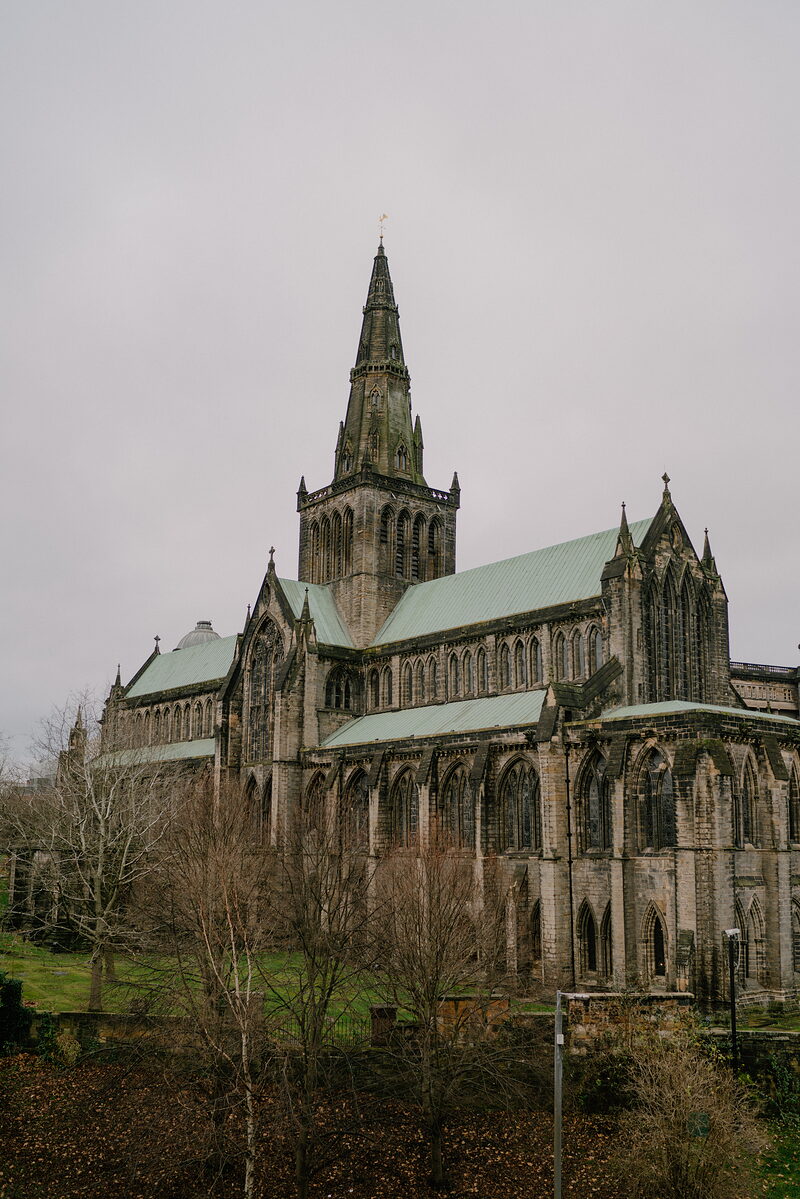 A gothic cathedral's tall crowned spire and green copper roofs rise above bare trees, tracery windows lining the nave below.