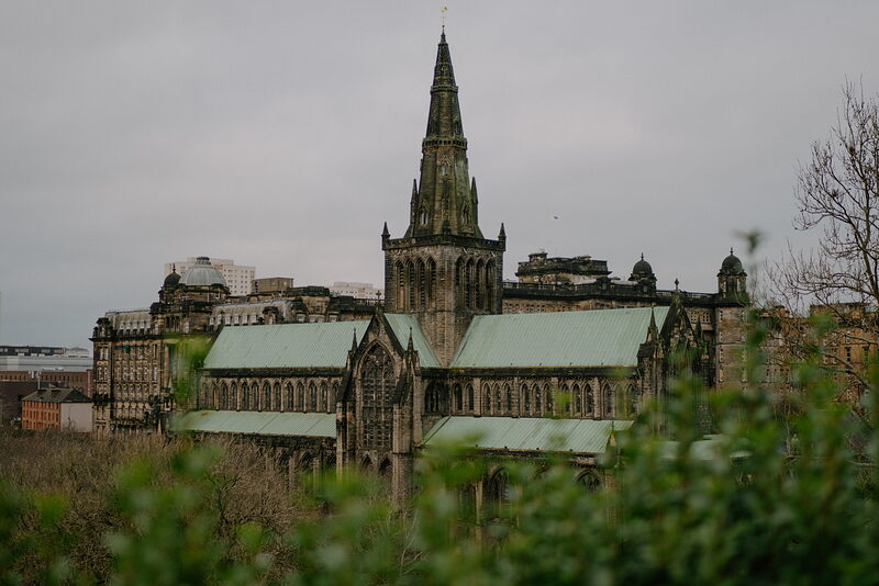 A gothic cathedral's spire and green copper roofs rise above the city, foliage framing the view in the foreground.
