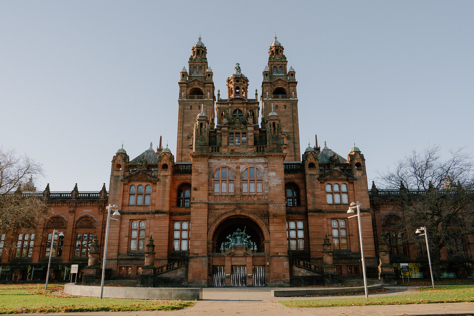 A red sandstone gallery building with twin spired towers and ornate pinnacles rises above a lawn and lamp posts.