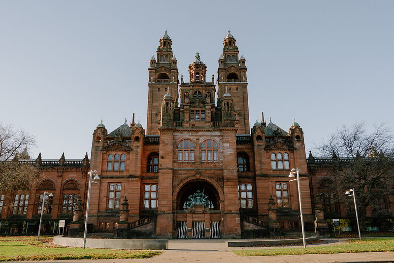 A red sandstone gallery building with twin spired towers and ornate pinnacles rises above a lawn and lamp posts.