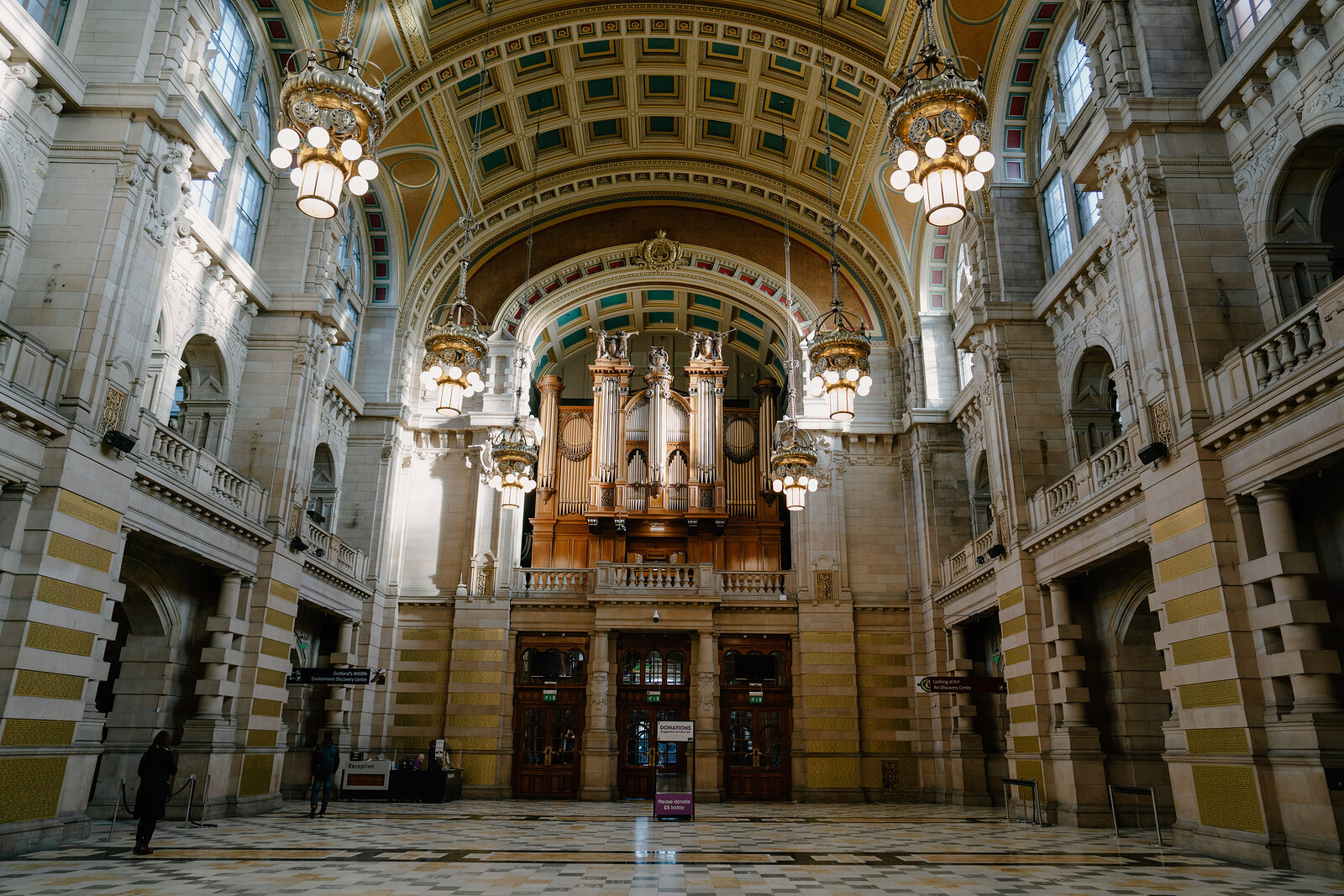 Ornate chandeliers hang beneath a vaulted, painted ceiling in a grand museum hall, a decorated pipe organ built into the far wall.