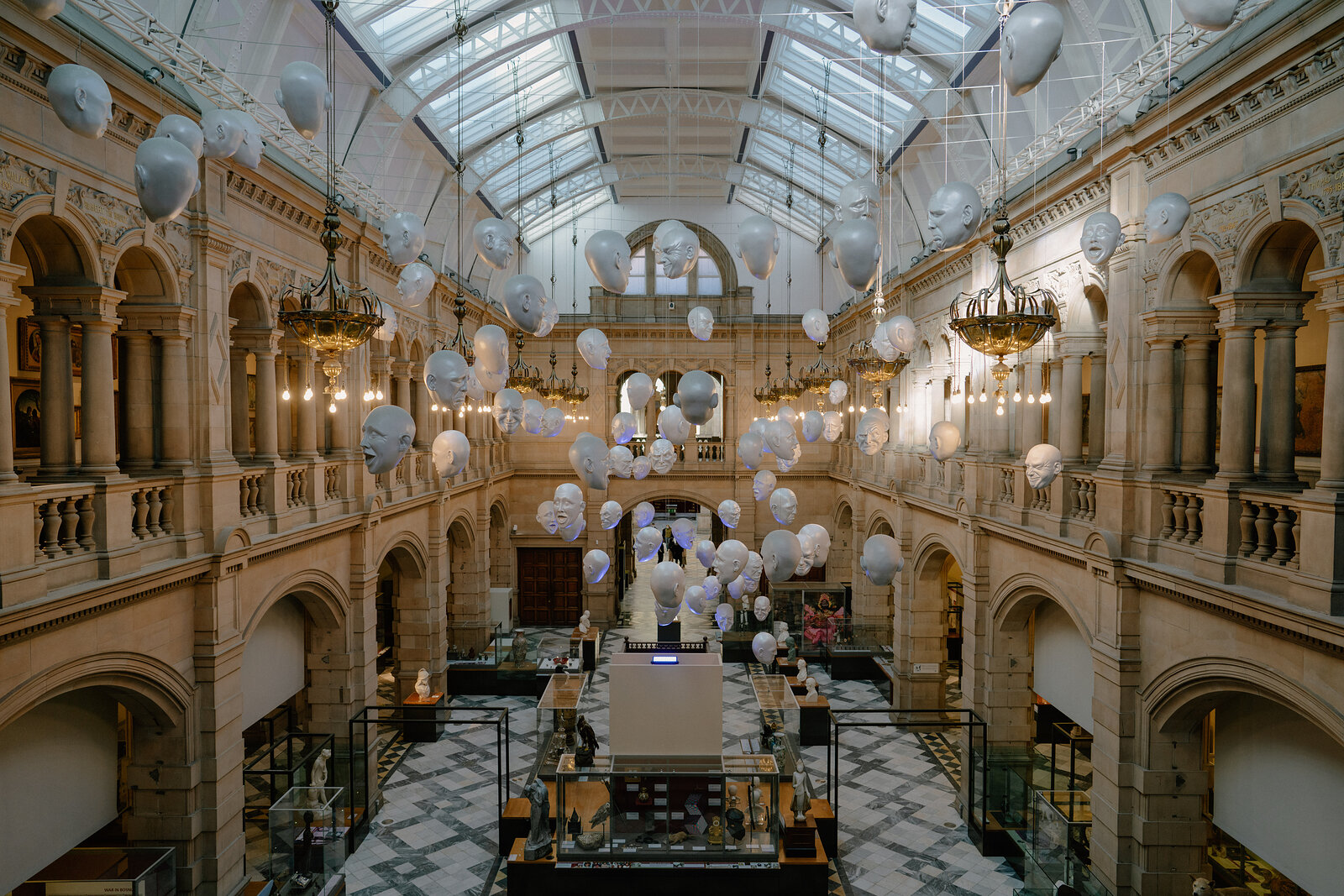 Rows of pale sculpted heads hang suspended beneath a glazed roof in a grand museum hall, an information stand set on the checkerboard marble floor below.