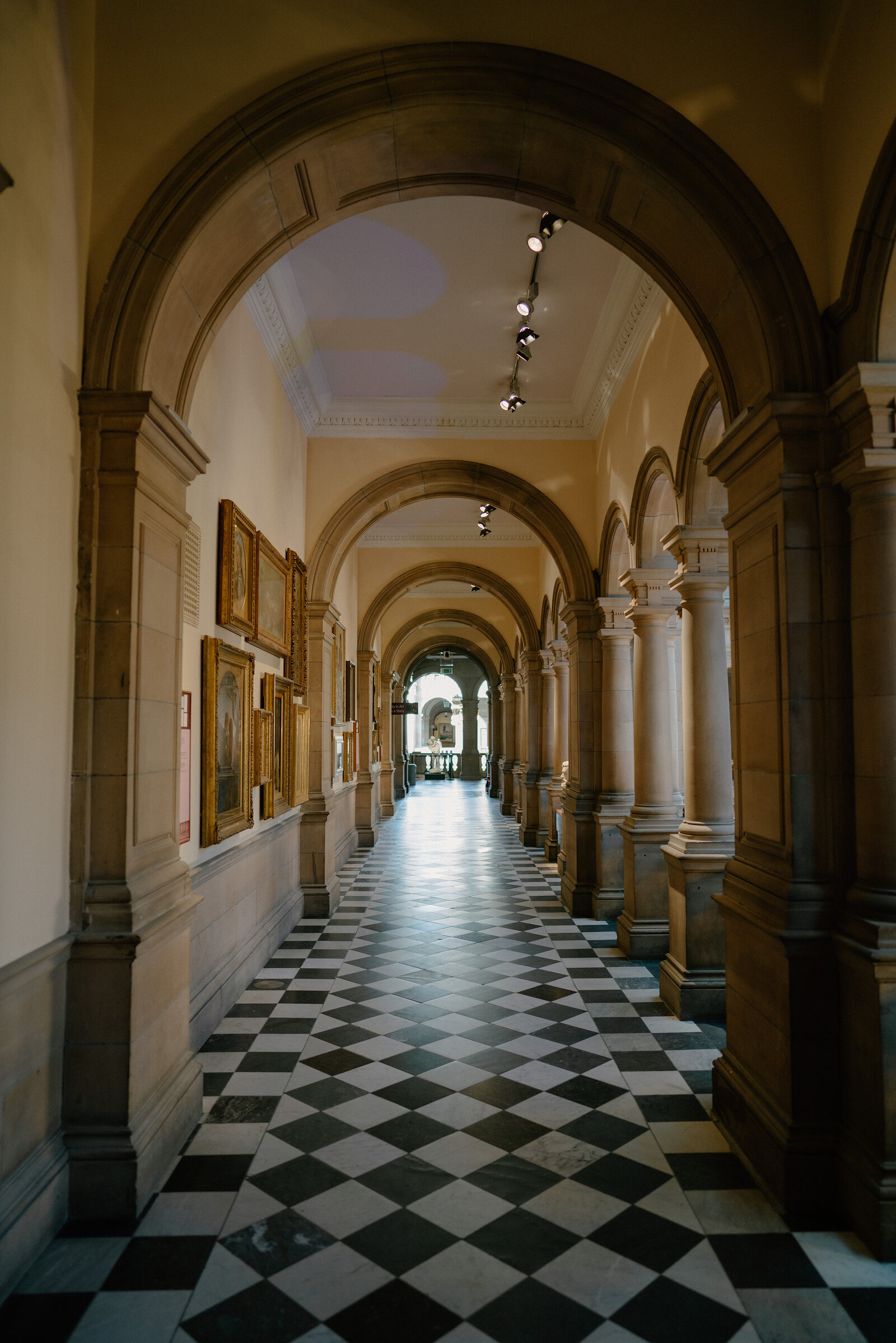 A vaulted stone corridor lined with arched columns and framed paintings leads toward a distant courtyard, checkerboard tiles underfoot.