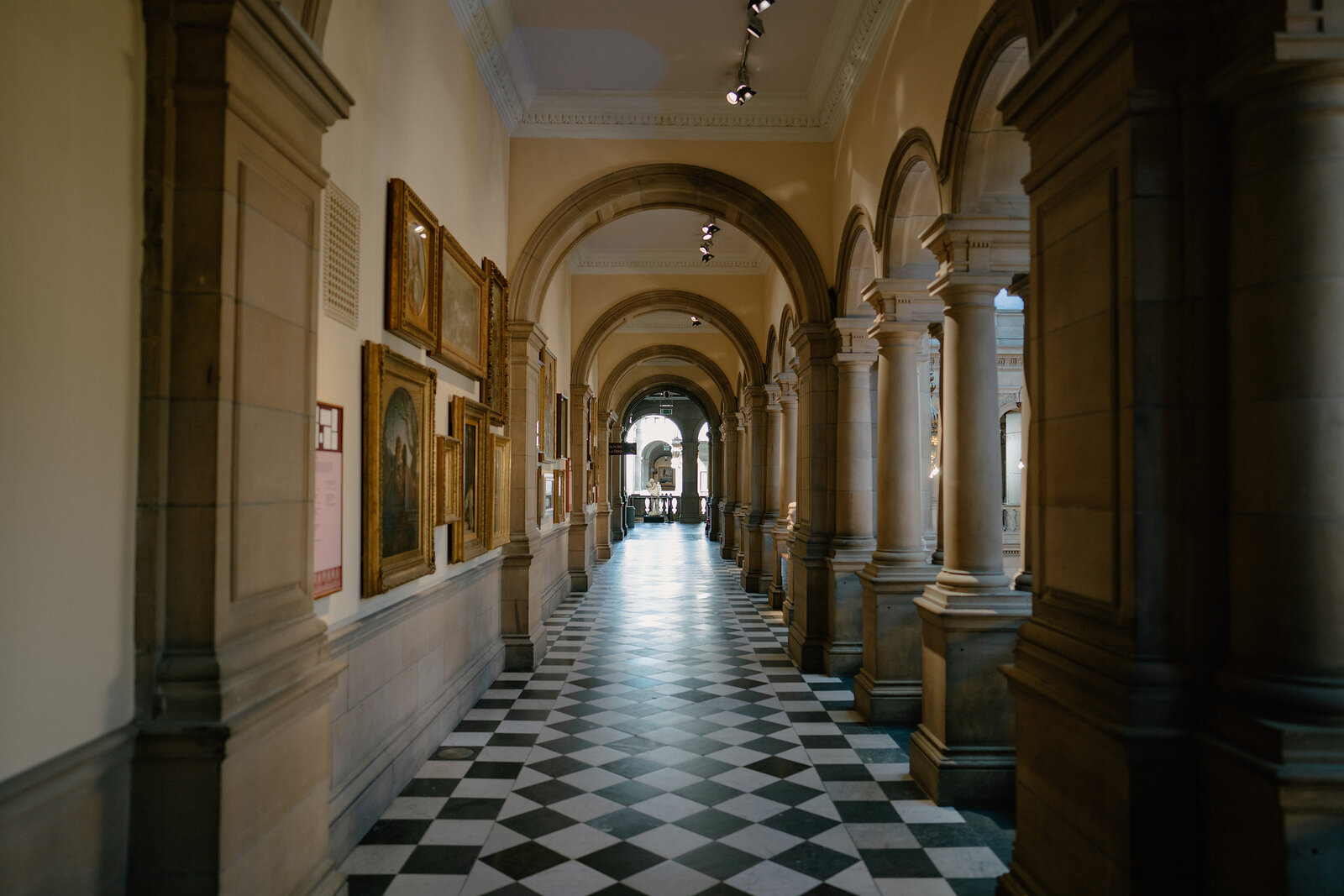 A vaulted stone corridor lined with arched columns and framed paintings leads past a distant doorway, checkerboard tiles underfoot.