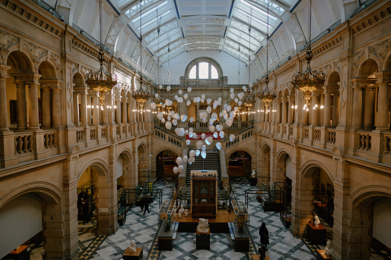 Rows of pale sculpted heads hang suspended beneath a glazed roof in a grand museum hall, visitors seated at tables on the marble floor below.