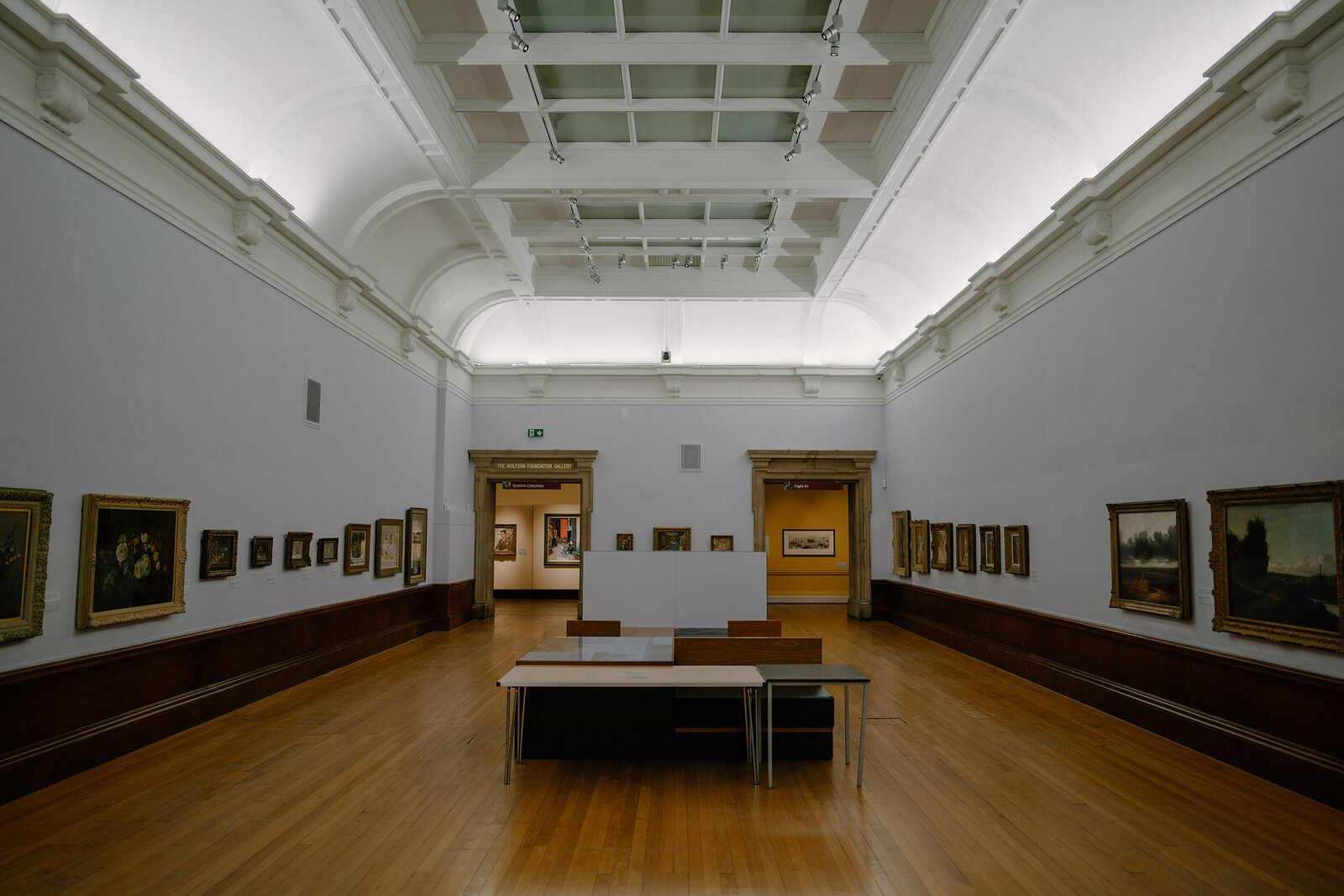 A long gallery room with a vaulted skylight ceiling displays rows of framed paintings along both walls, a bench set in the centre of the wooden floor.