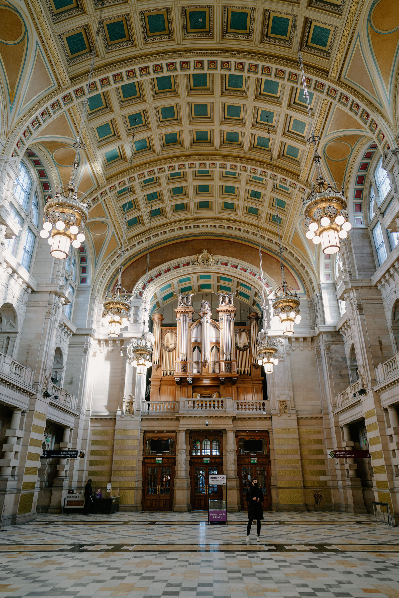 A vaulted, painted ceiling with gilded panels rises above a decorated pipe organ built into a grand museum hall, chandeliers hanging beneath the arches.