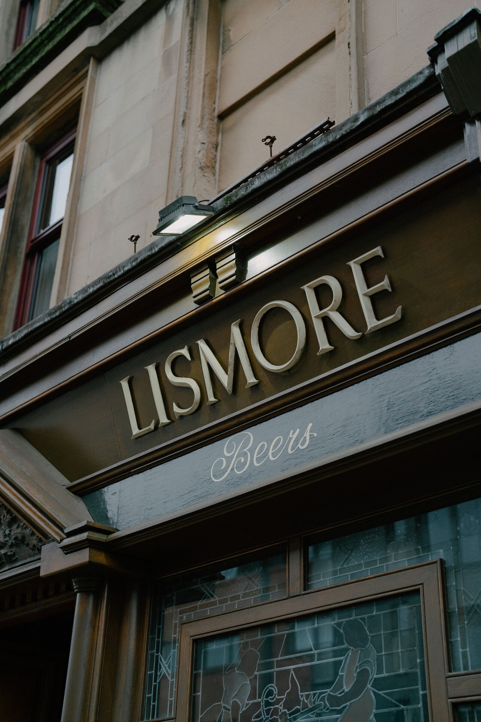 A gold-lettered sign reading 'Lismore, Beers' spans the corner of a sandstone pub building, stained-glass windows below.
