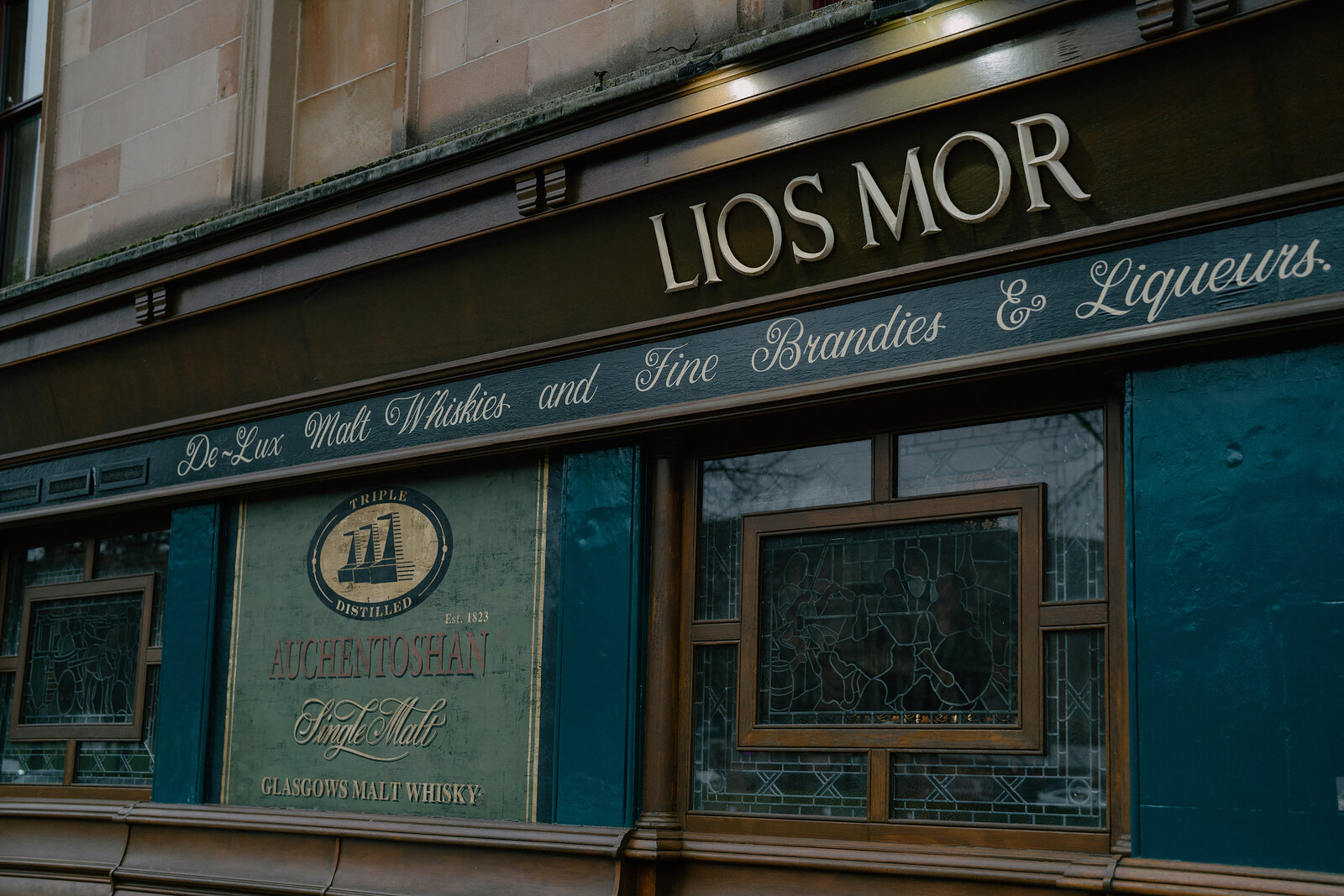 A green shopfront carries gold lettering reading 'Lios Mor, De-Lux Malt Whiskies and Fine Brandies & Liqueurs' above stained-glass windows.