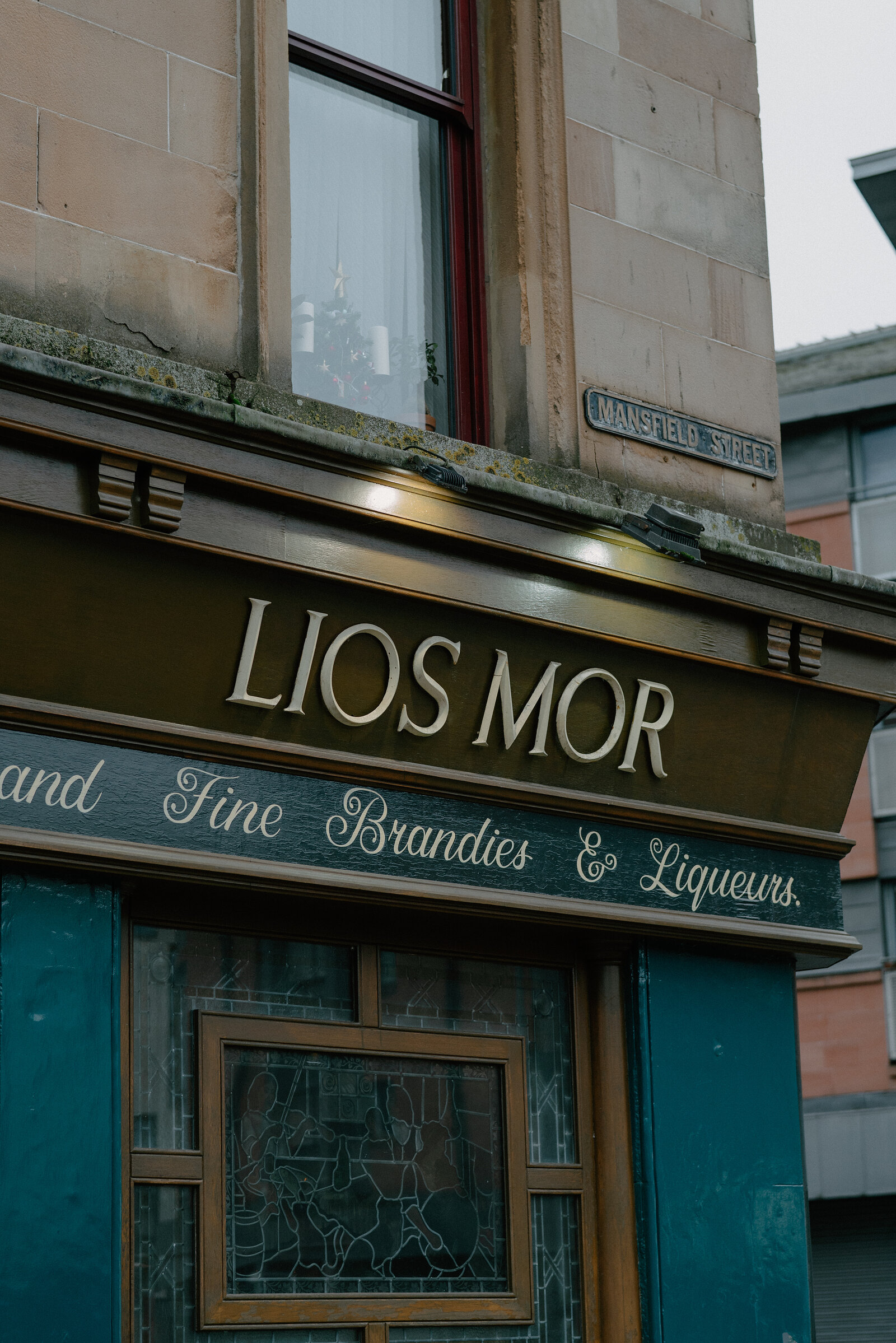 A gold sign reading 'Lios Mor' and 'Fine Brandies & Liqueurs' spans a sandstone shopfront beside a street sign for 'Mansfield Street'.