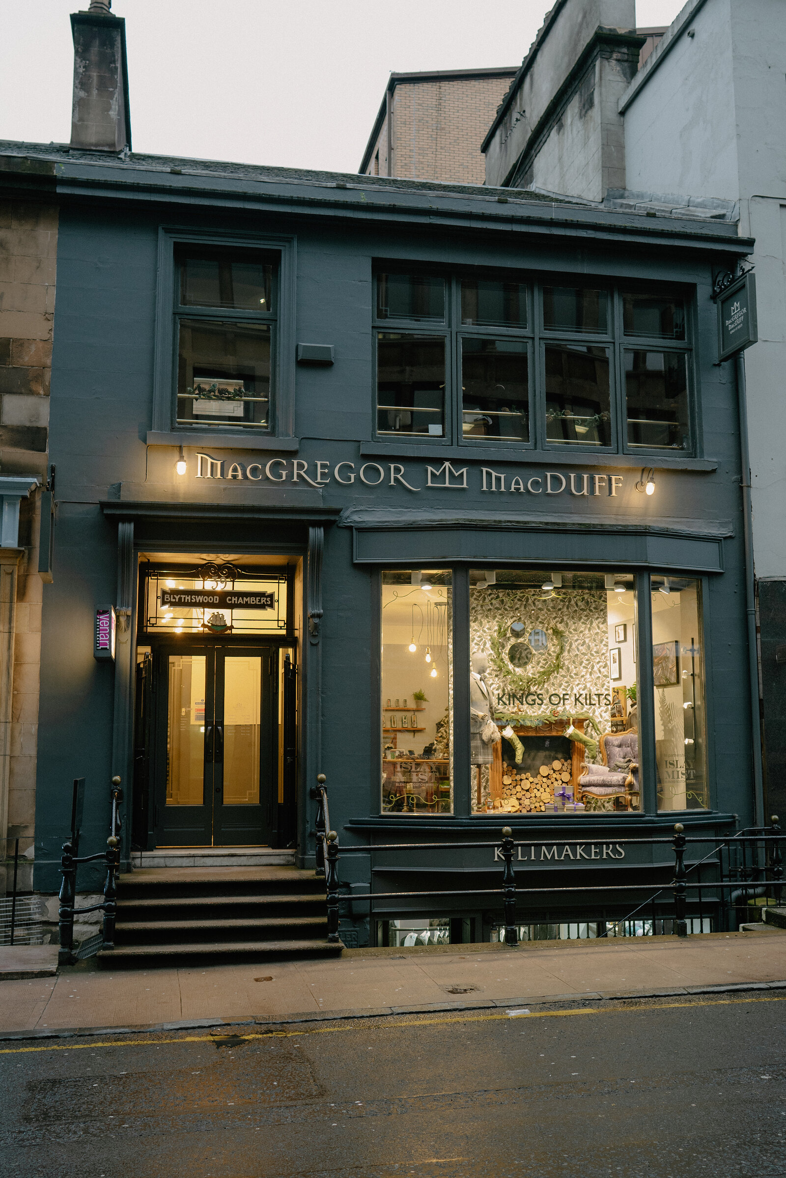 A dark green shopfront carries a sign reading 'MacGregor MacDuff, Kings of Kilts, Kiltmakers', a decorated window display visible beside the entrance.