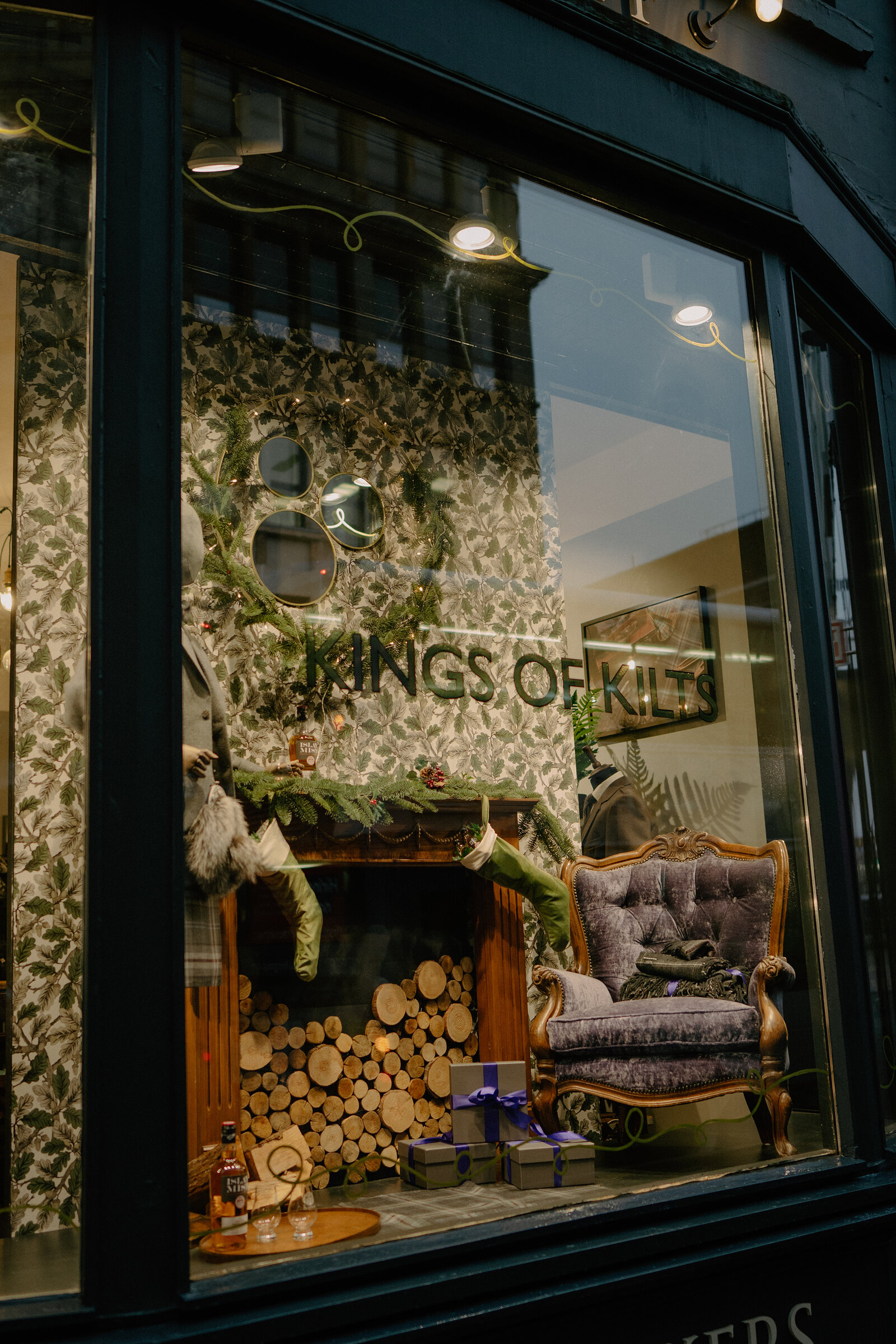 A shop window display reads 'Kings of Kilts' above a fireplace stacked with logs, a tartan-draped armchair and wrapped gifts arranged beside it.