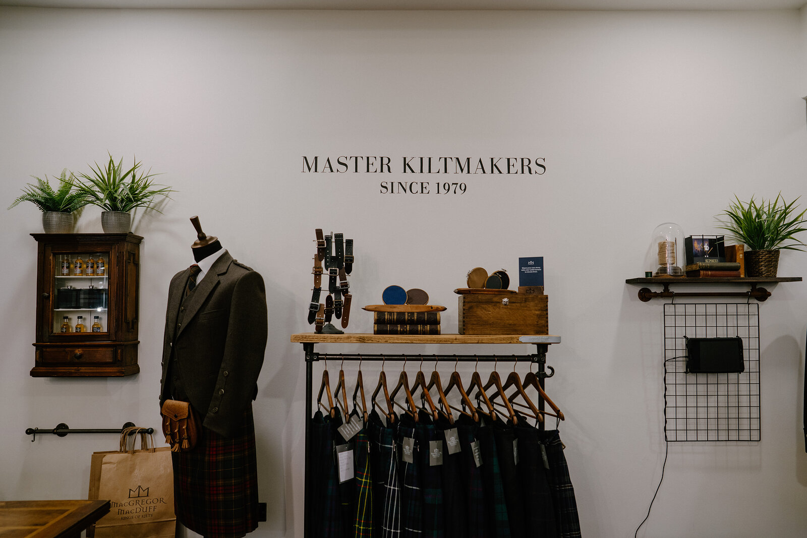 A wall reads 'Master Kiltmakers, Since 1979' above a mannequin in a tweed jacket and kilt, hanging kilts and accessories displayed on shelving beside it.