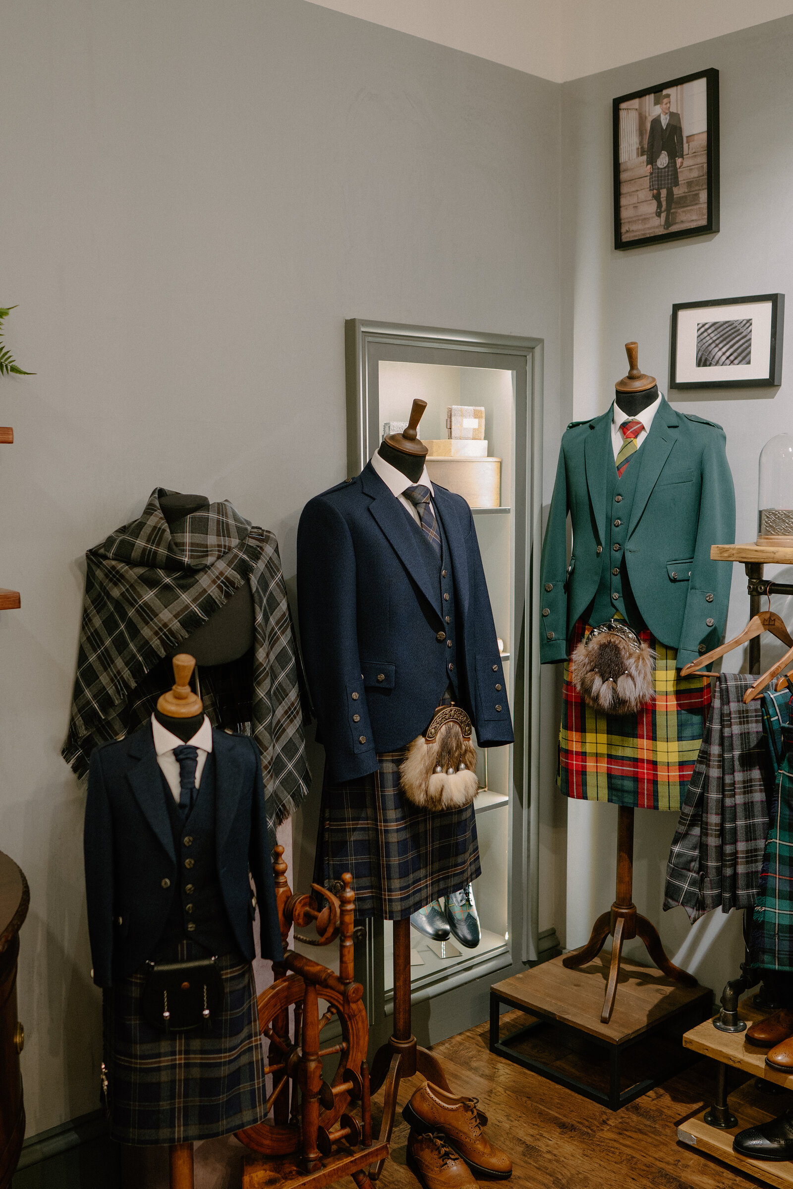 Mannequins in a shop display tartan kilt outfits with sporrans and tweed jackets, a green tweed waistcoat and jacket set beside a mirror.