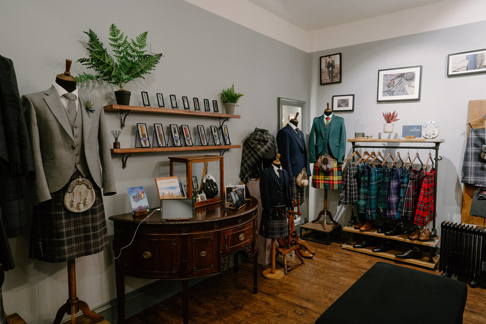 A shop displays tweed jackets and tartan kilts on mannequins and hanging rails beside an antique sideboard and framed photographs.