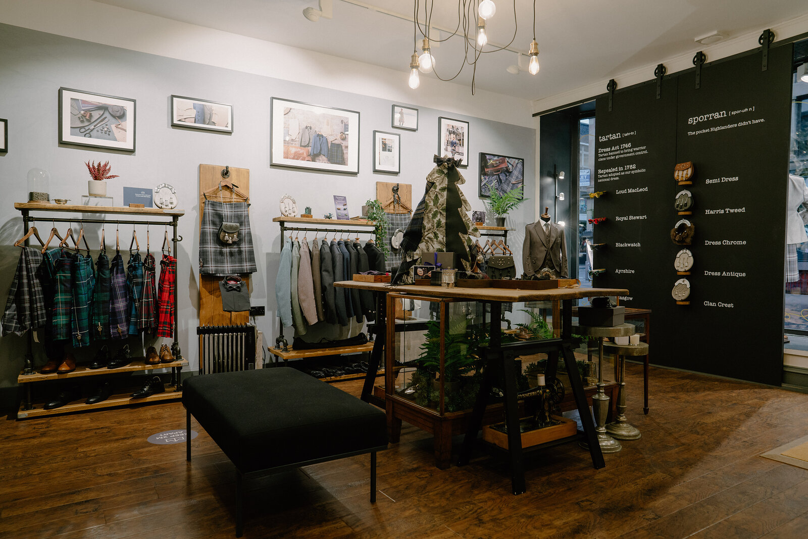 A shop displays checked shirts on hanging rails beside a small Christmas tree and framed art prints, a sign listing product details on the wall.