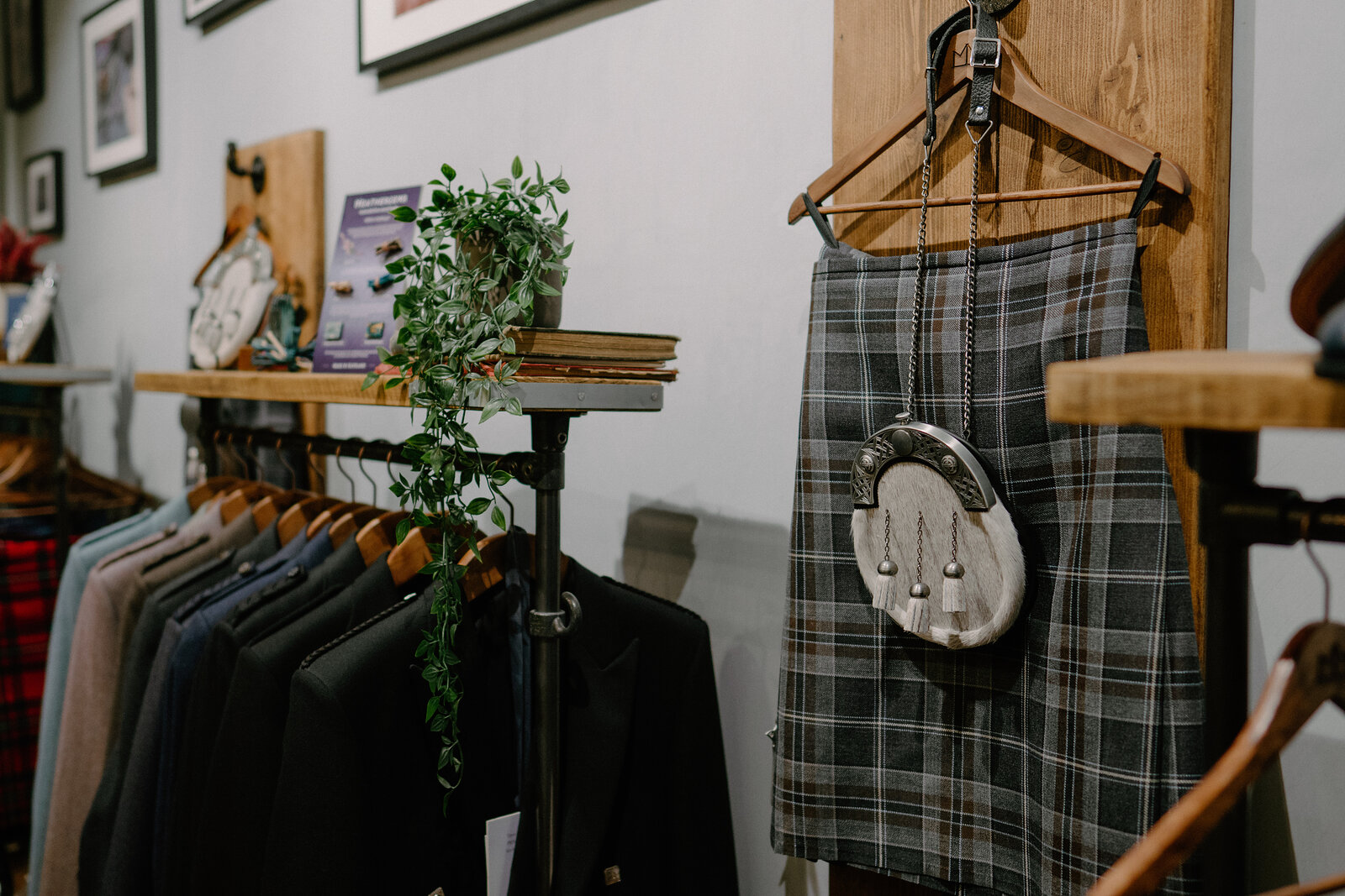 A tartan kilt with a sporran hangs from a wooden hanger beside a rail of jackets, framed prints and trailing ivy on a shelf above.