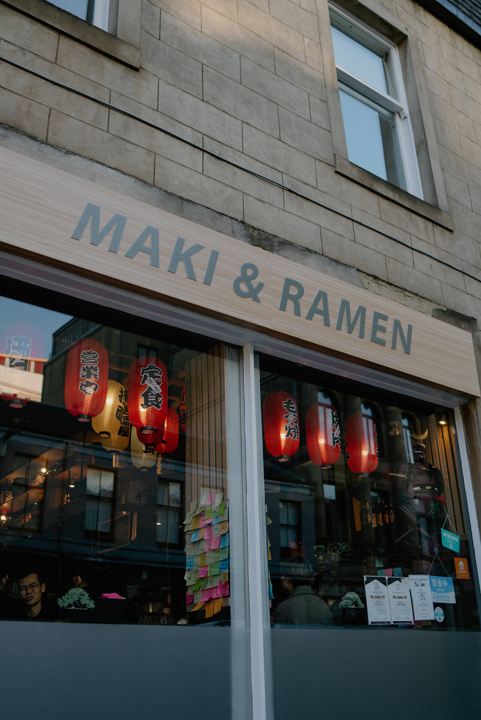 A sandstone shopfront carries a sign reading 'Maki & Ramen' beside a window strung with red paper lanterns, diners visible inside.