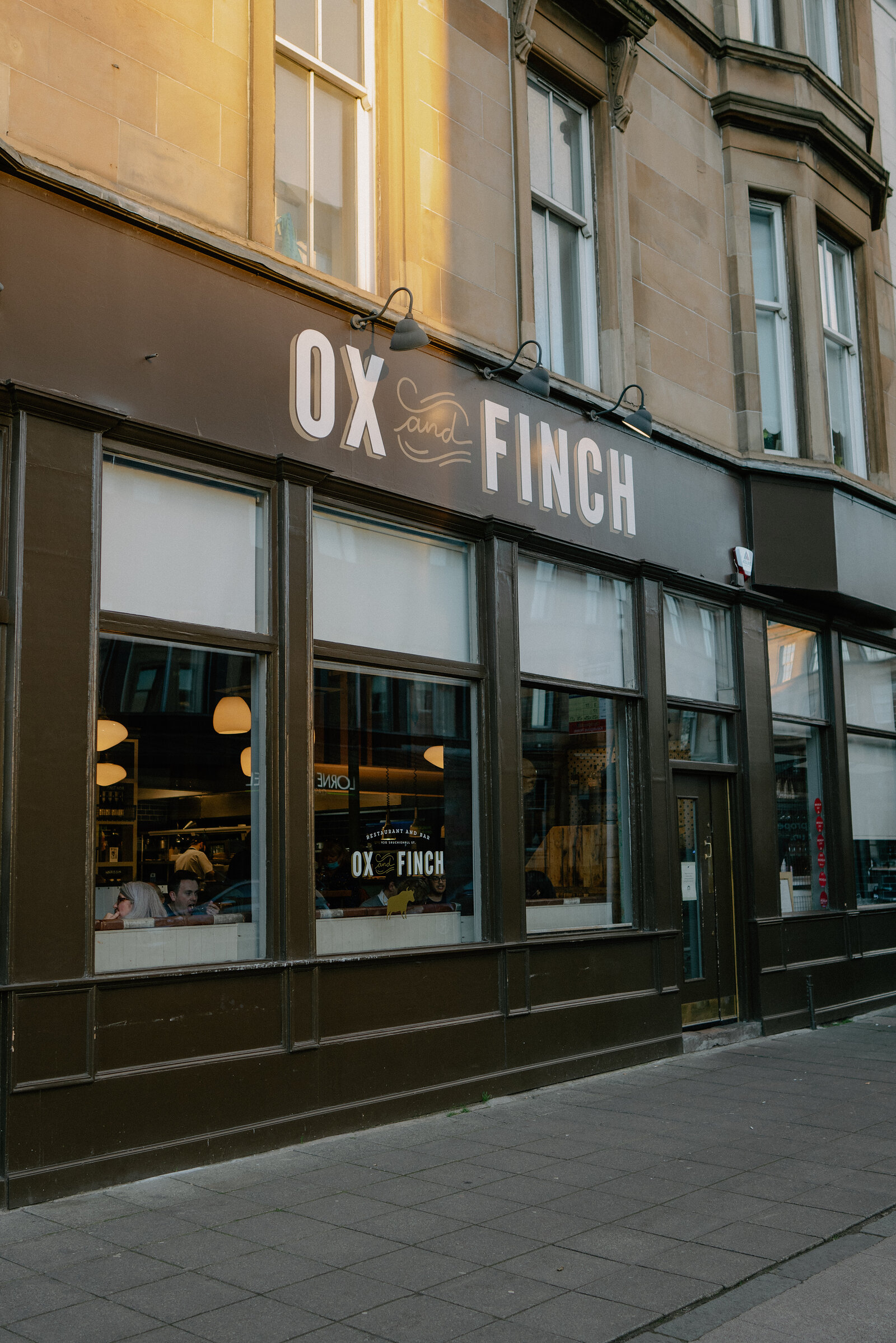 A dark shopfront carries a sign reading 'Ox and Finch' above tall windows showing diners seated inside, sconce lights mounted along the facade.