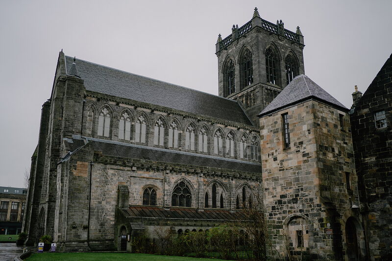 A large sandstone church with a central tower, traceried windows and an arcaded cloister stands beside a wet lawn.