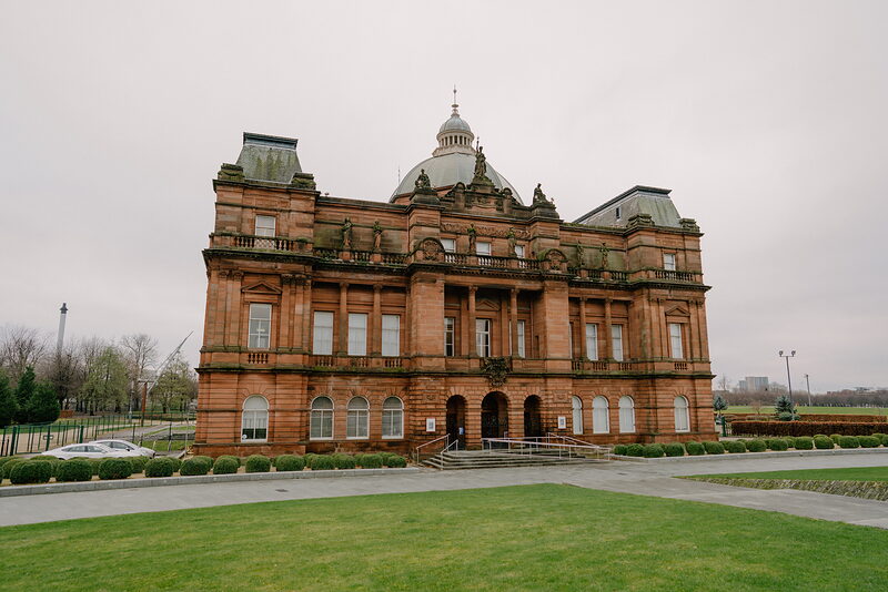 A grand red sandstone building with a central dome and pilastered facade stands behind a formal lawn and paved paths.