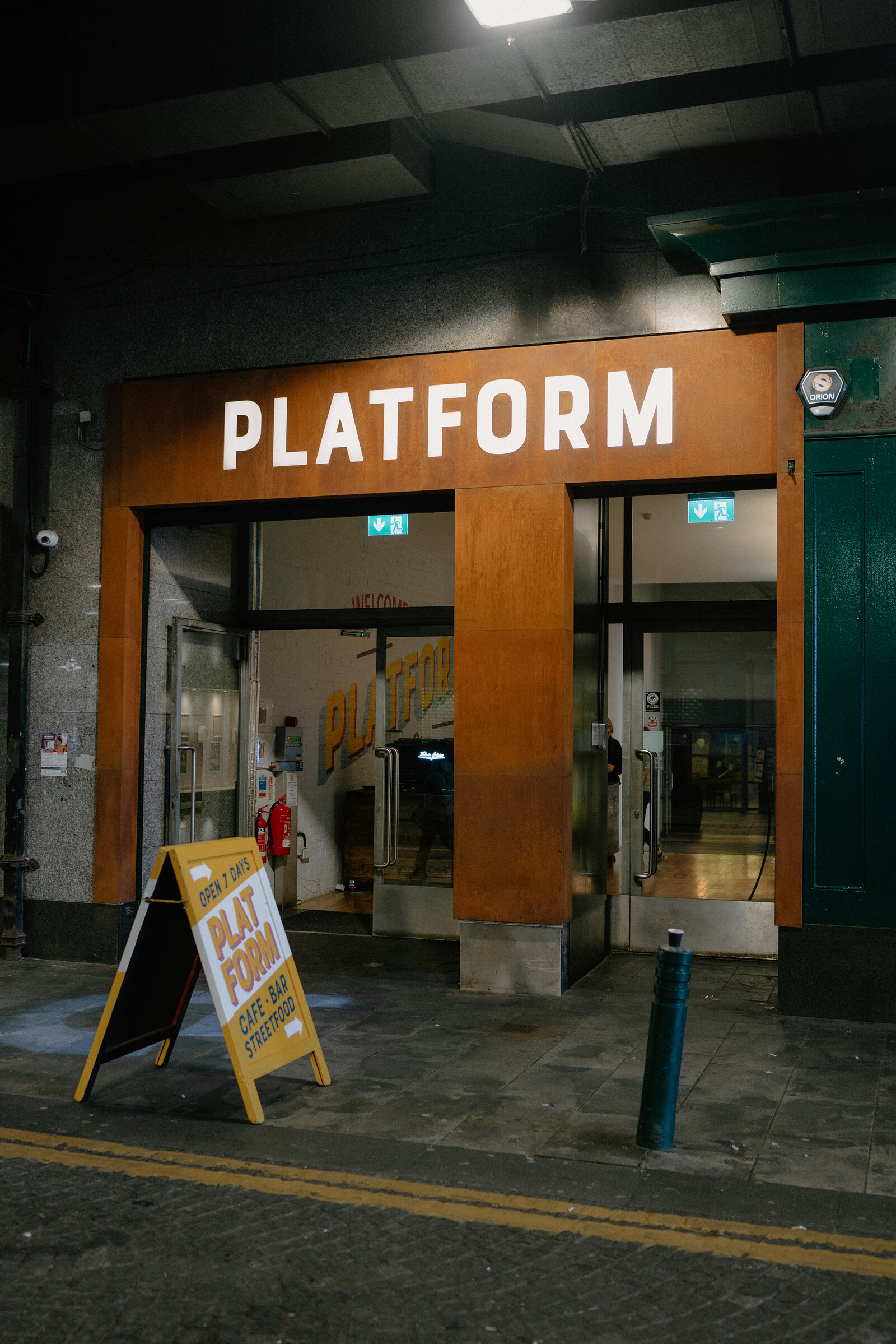 A lit café bar entrance carries a large sign reading 'Platform' beneath a railway arch, a sandwich board and bollards standing on the pavement outside.