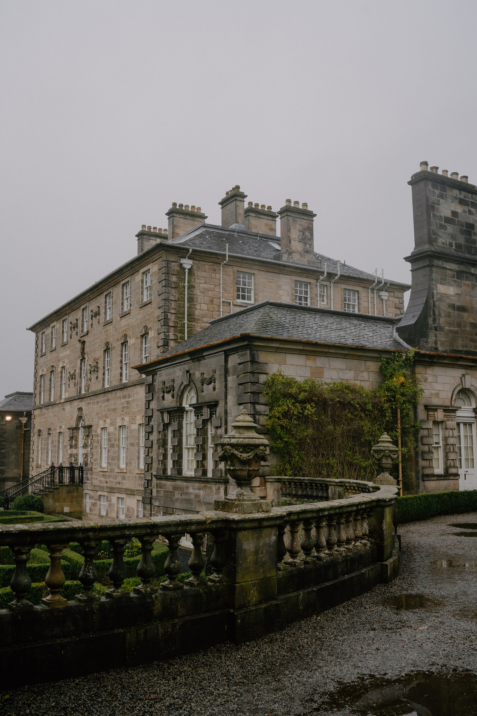 A grand stone mansion with multiple chimney stacks and ivy-covered walls stands behind a balustraded terrace and clipped hedges.
