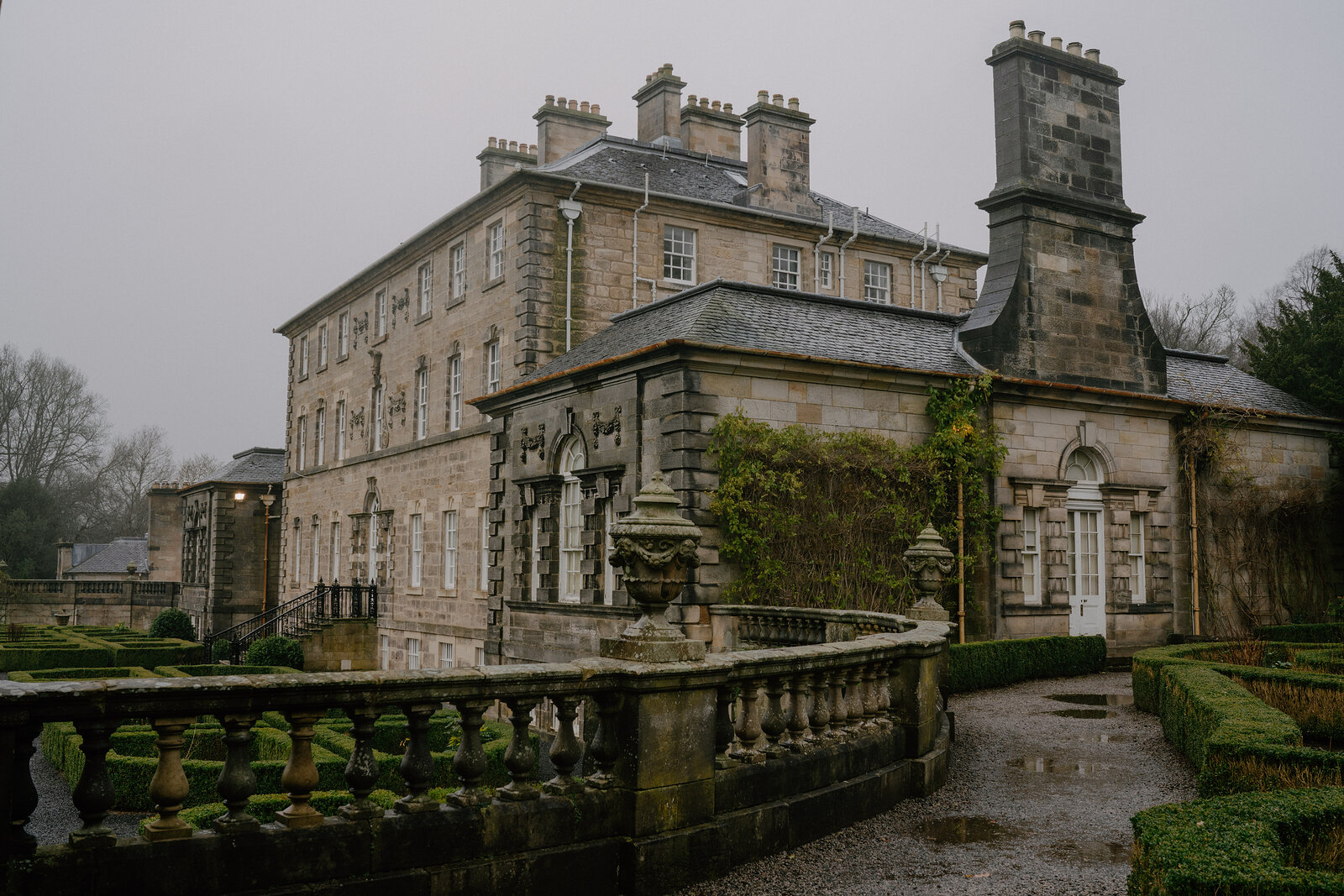 A grand stone mansion with a carved porch and balustraded terrace stands behind clipped box hedges in a formal garden.