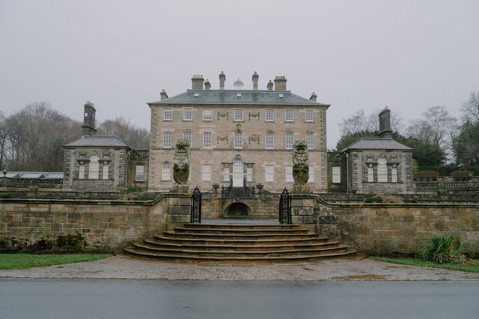 A grand stone mansion with a symmetrical facade and carved urns stands behind a curved flight of steps and gate piers.