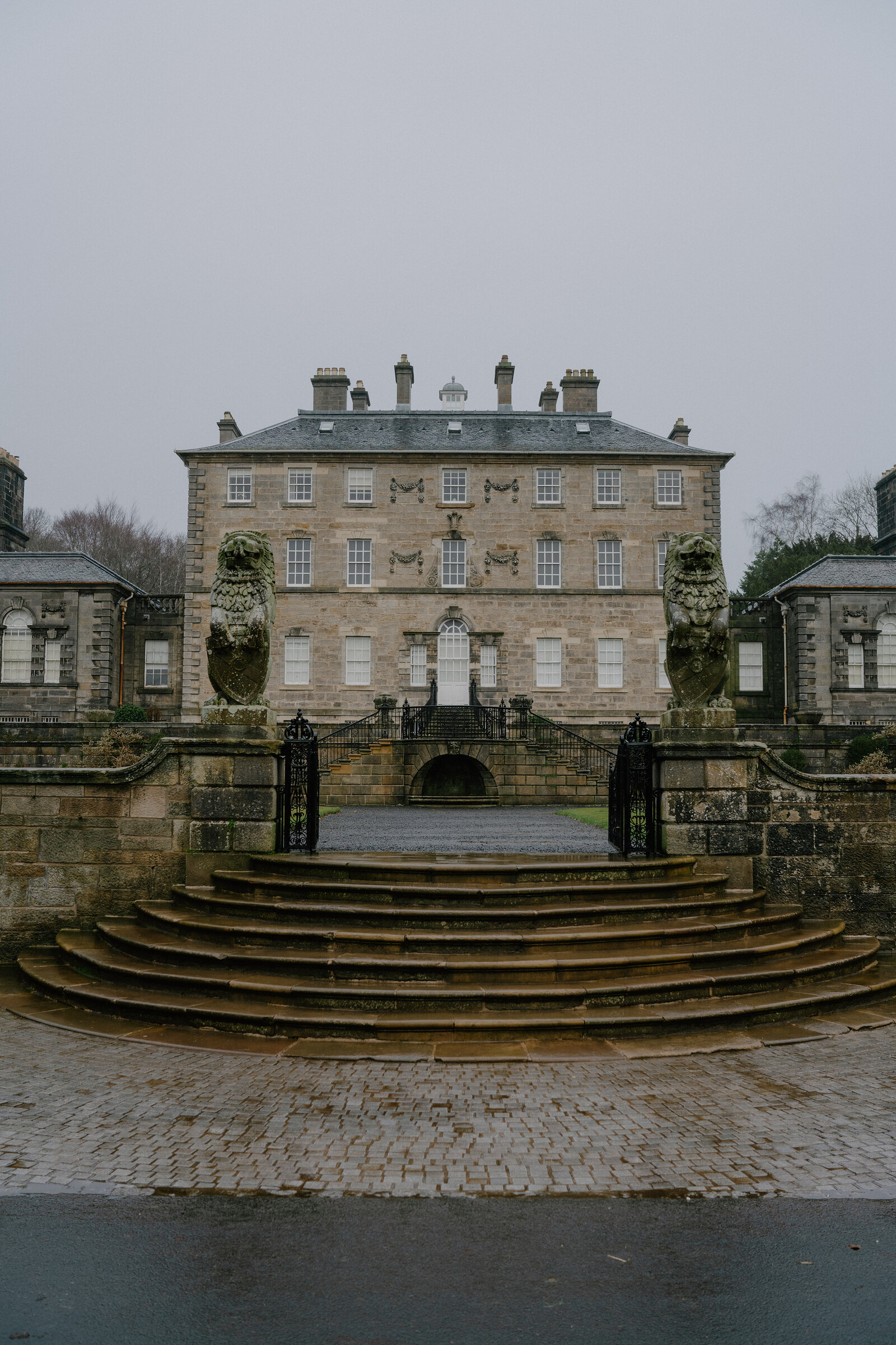 A grand stone mansion with a symmetrical facade and carved urns stands behind a curved flight of steps and cobbled forecourt.