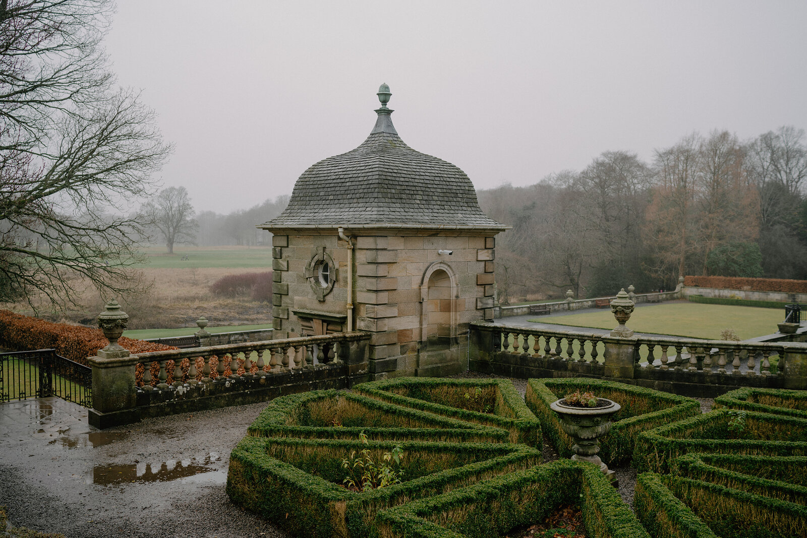 An octagonal stone gazebo with a domed roof stands at the corner of a formal garden, box hedges arranged in a knot pattern below.