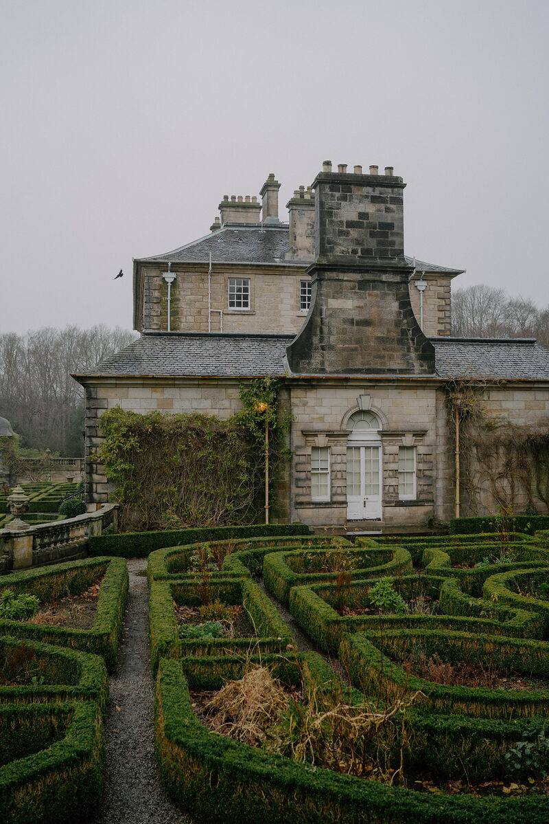 A stone mansion with tall chimney stacks and ivy-covered walls overlooks a formal box-hedge parterre garden.