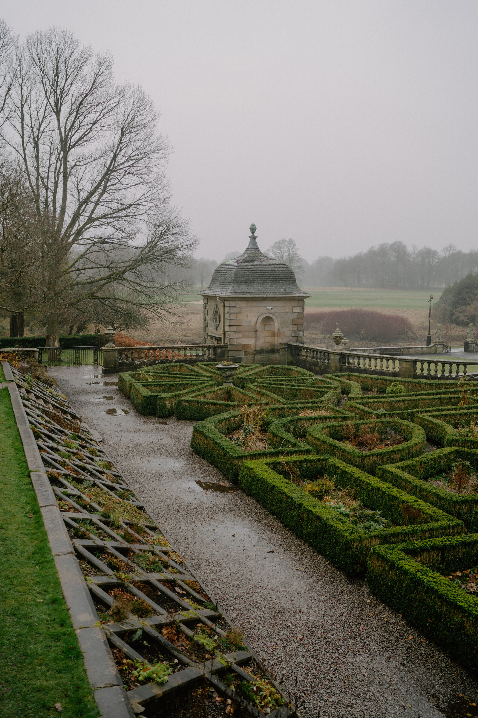 A gravel path winds through a formal box-hedge garden toward a stone gazebo with a domed roof, misty parkland stretching beyond a balustrade.