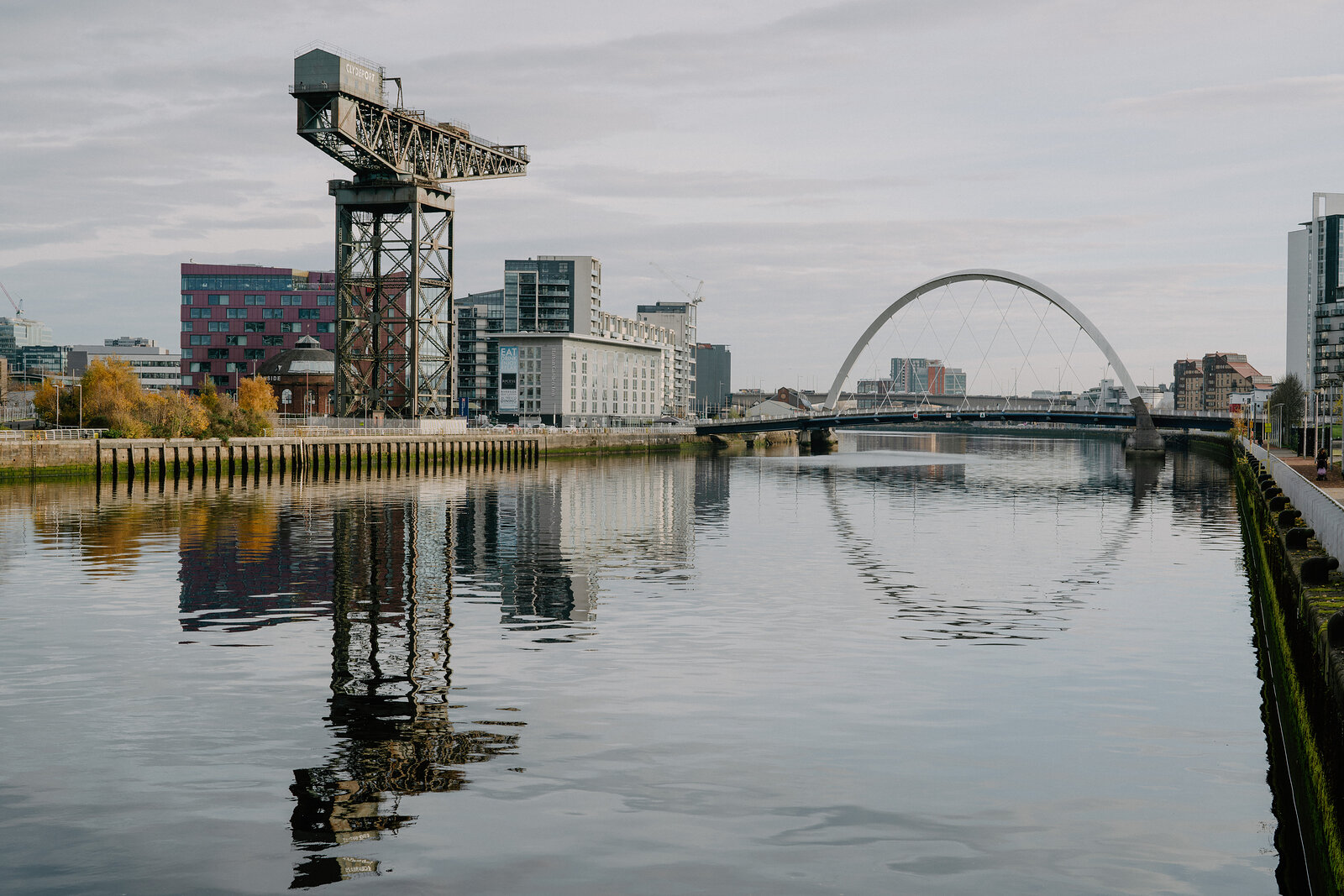 A tall black cantilever crane stands on a riverbank beside a modern glass hotel, a white arched footbridge visible further along the water.