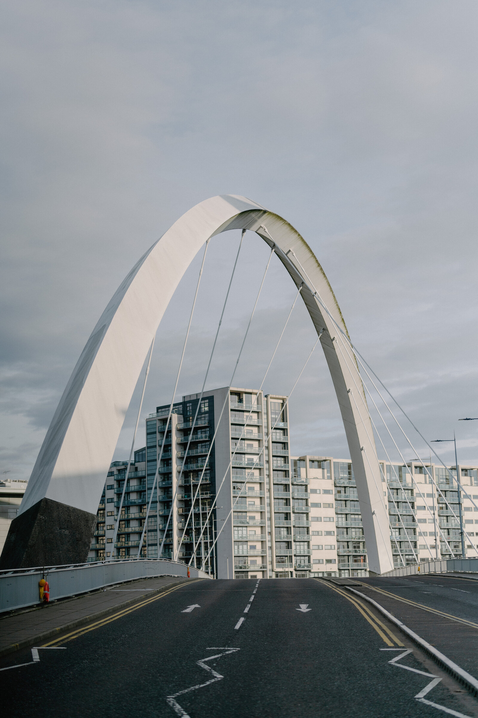 A white bowed pedestrian bridge with angled struts and cable stays rises against a cloudy sky above a road, apartment buildings visible behind.