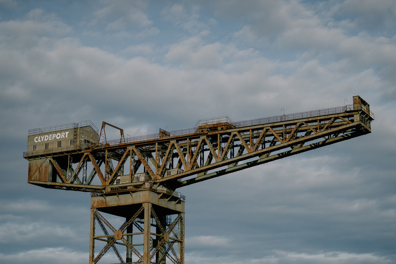 A rusted cantilever crane stamped 'Clydeport' rises against a cloudy sky beside a riverside walkway.
