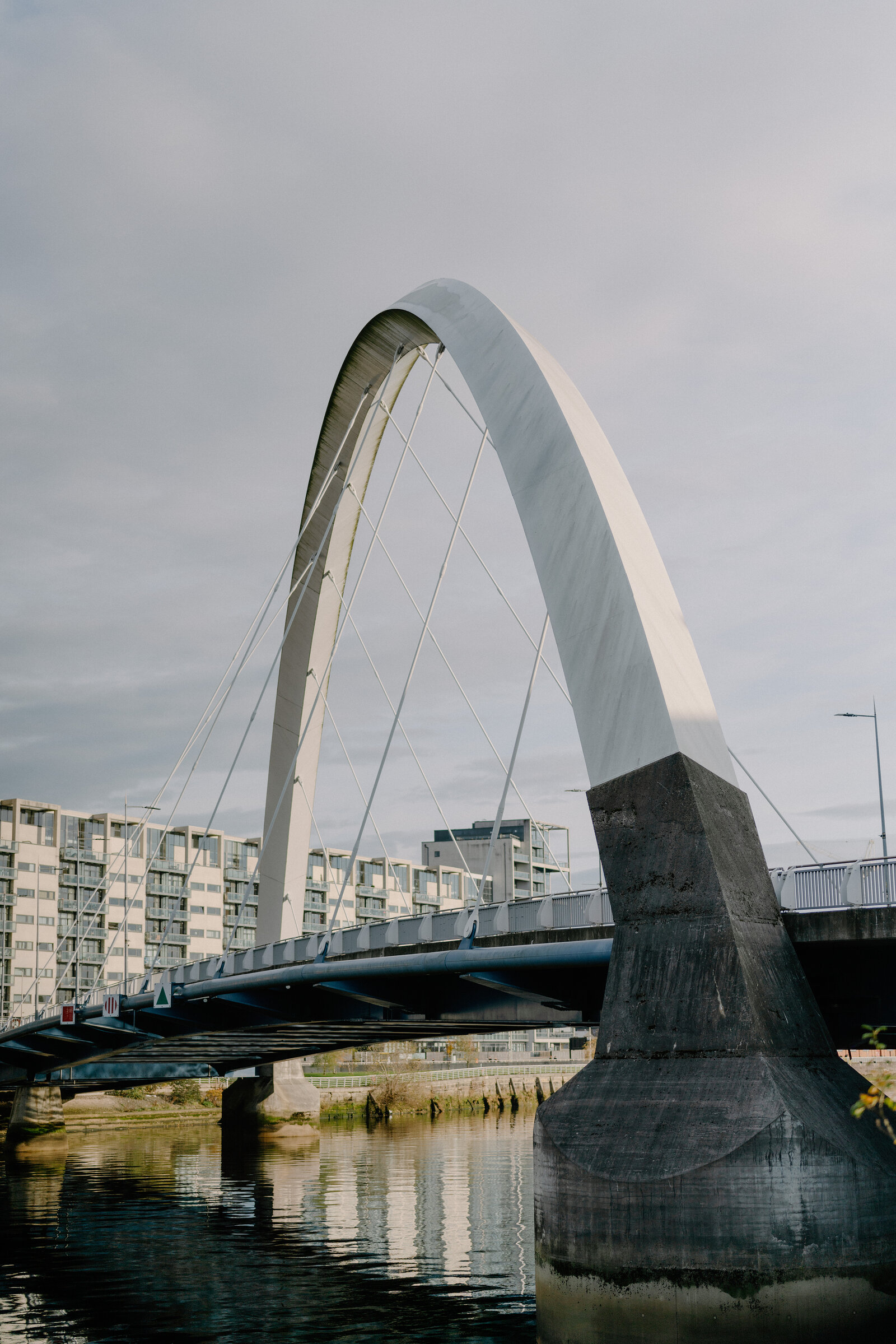 A white bowed pedestrian bridge with angled struts and cable stays spans a calm river beside a stone pier, apartment buildings reflected in the water.