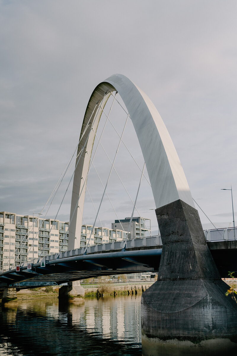 A white bowed pedestrian bridge with angled struts and cable stays spans a calm river beside a stone pier, apartment buildings reflected in the water.