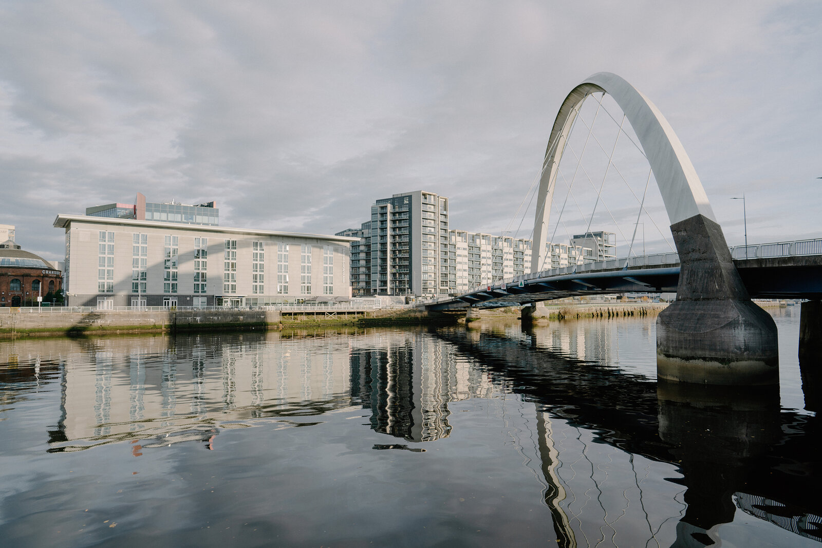 A white bowed pedestrian bridge with angled struts spans a calm river beside modern apartment buildings, reflected in the water.