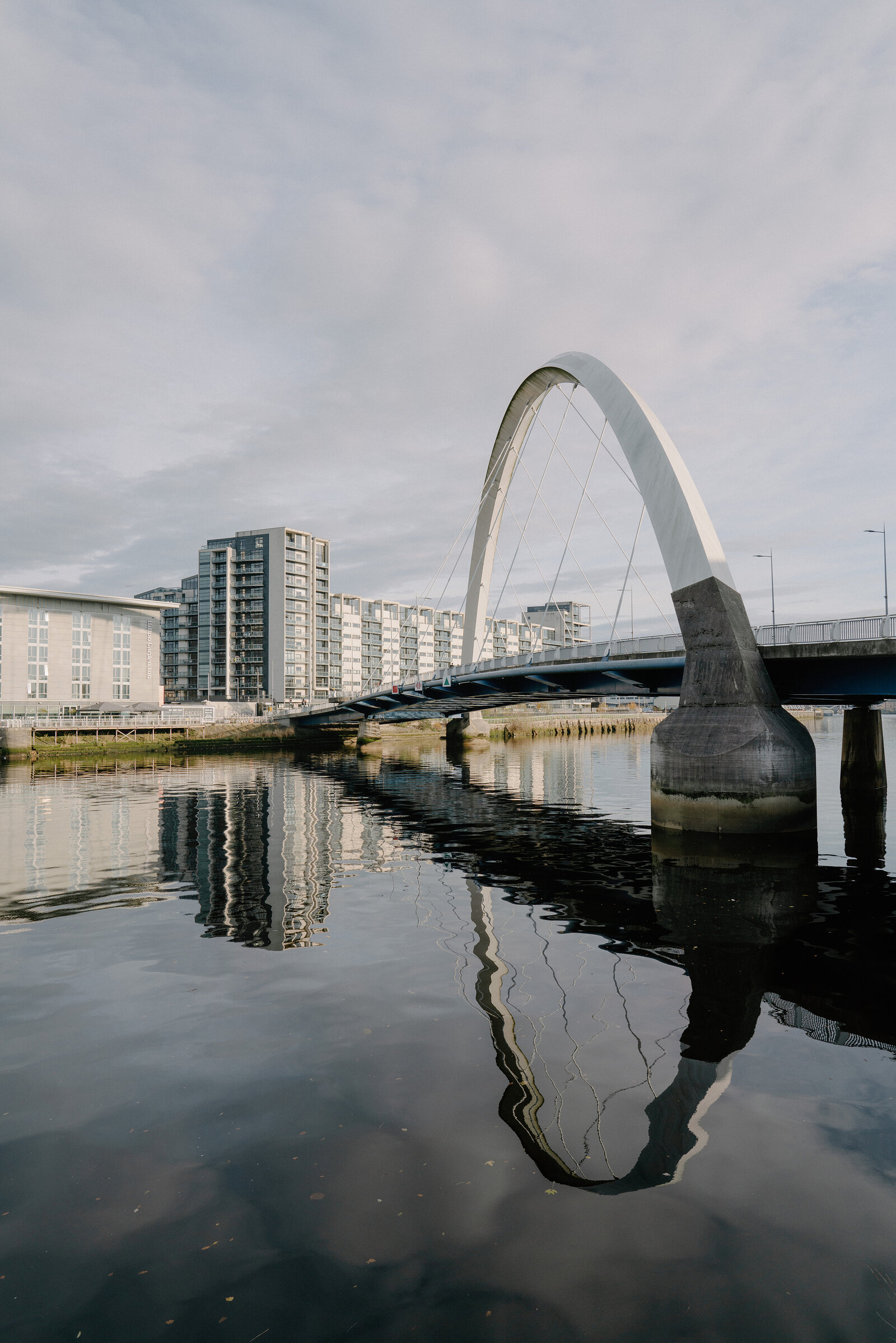 A white bowed pedestrian bridge with cable stays arches over a calm river beside modern apartment buildings, fully reflected in the water.
