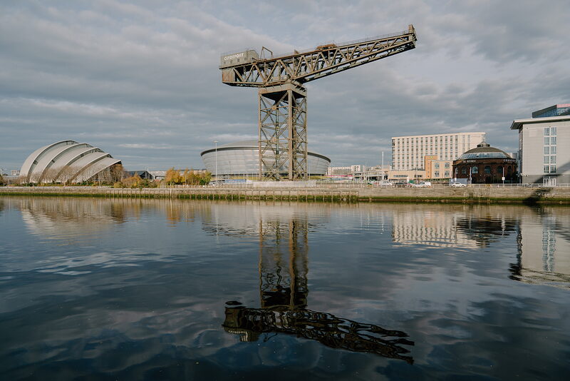 A rusted cantilever crane stands beside a curved silver-panelled auditorium and a triangular glass exhibition centre, all reflected in a calm river.