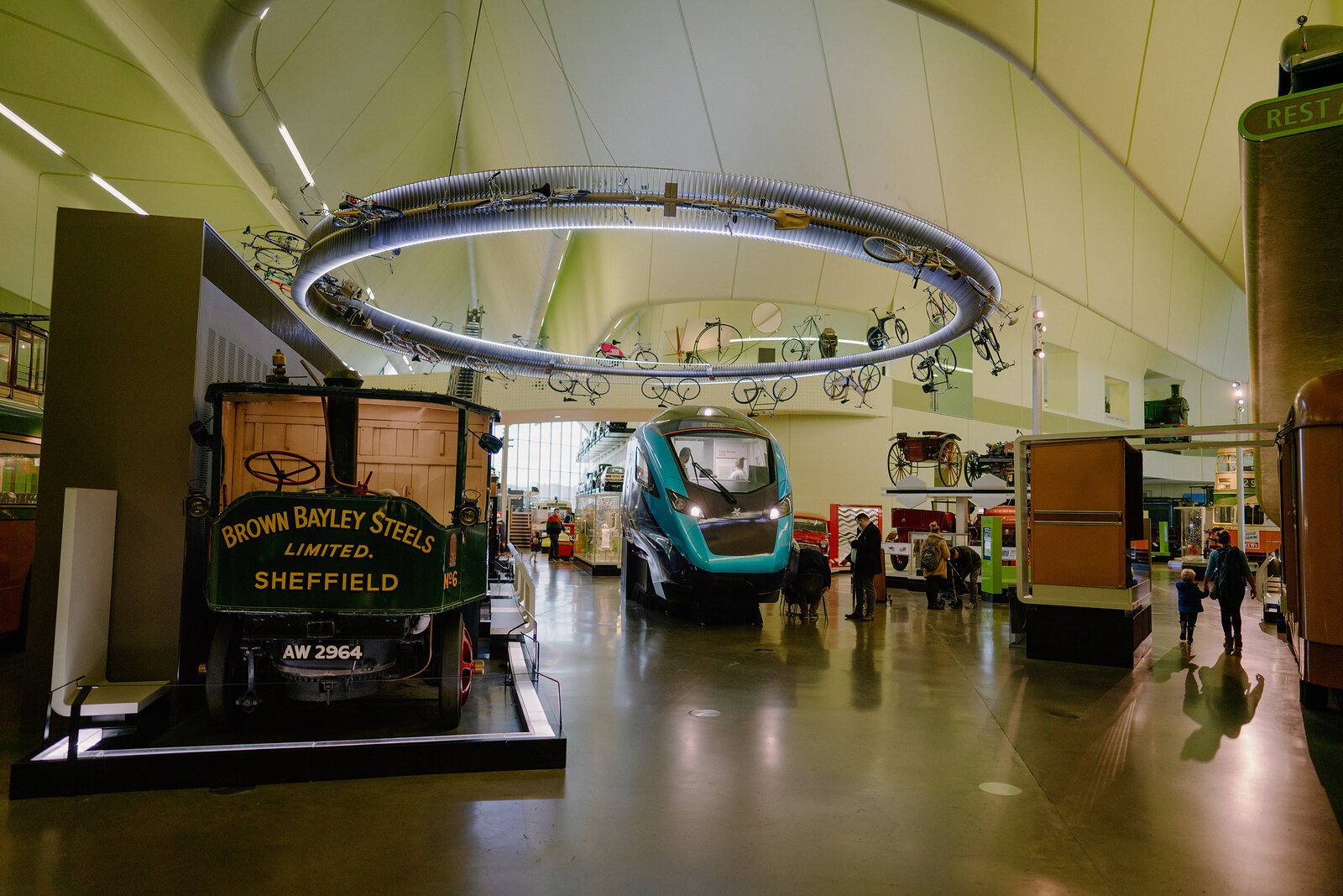 A museum gallery displays vintage vehicles including a horse-drawn cart lettered 'Brown Bayley Steels, Sheffield' beneath a circular ceiling track of hanging bicycles.
