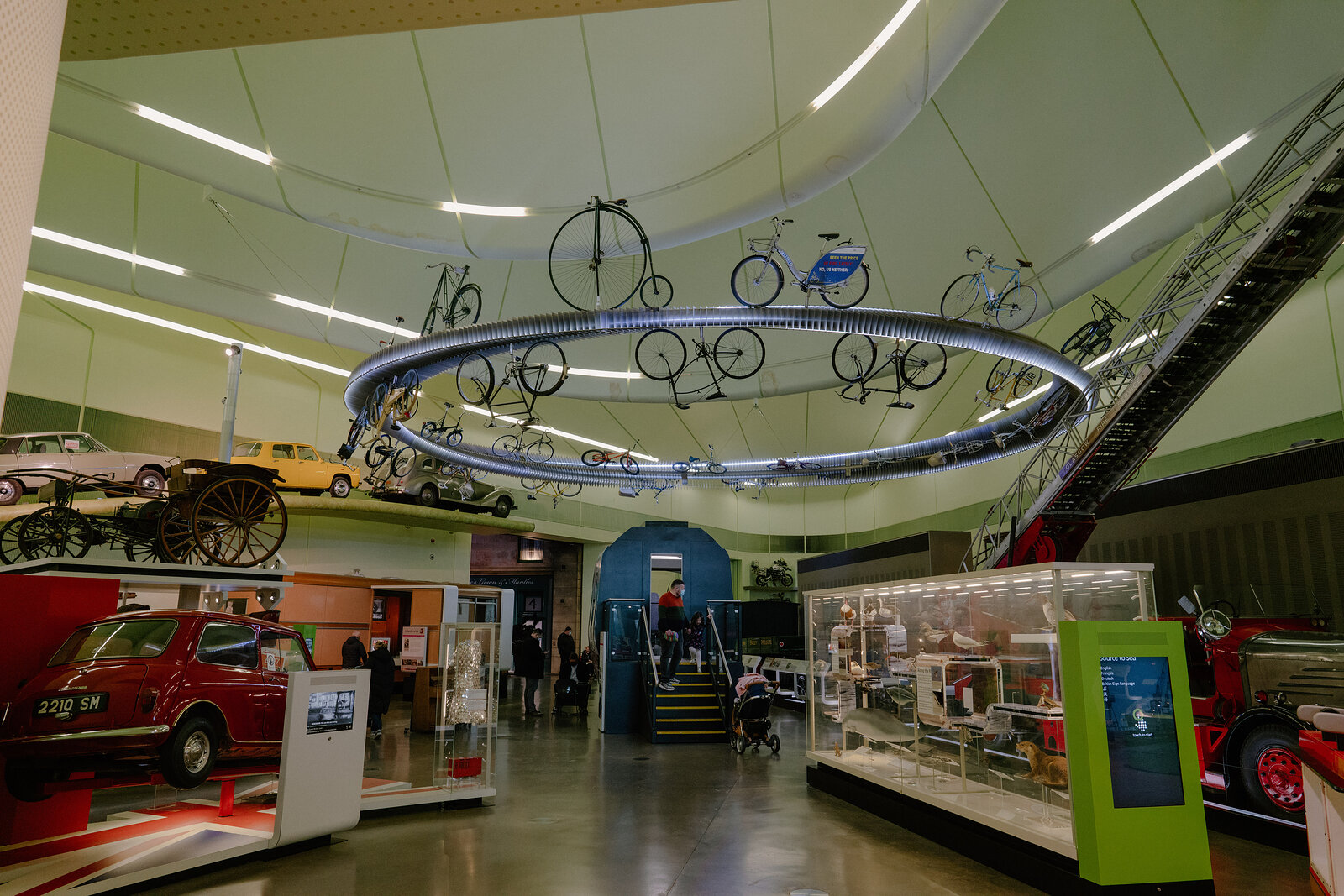 A museum gallery displays vintage vehicles including a red car and a fire engine beneath a circular ceiling track of hanging bicycles.